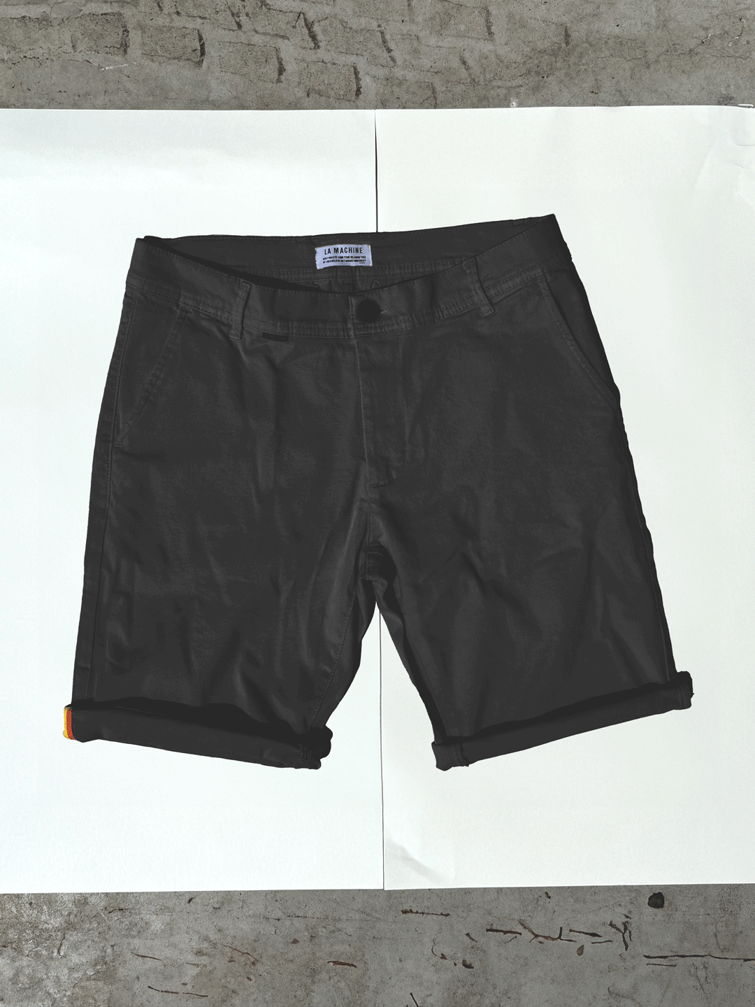 A pair of Grand Tours - Rest Day Bermuda shorts in black by La Machine Cycle Club with rolled cuffs and a semi-tailored fit is laid flat on a white background atop a concrete floor. The waistband features a visible clothing tag.