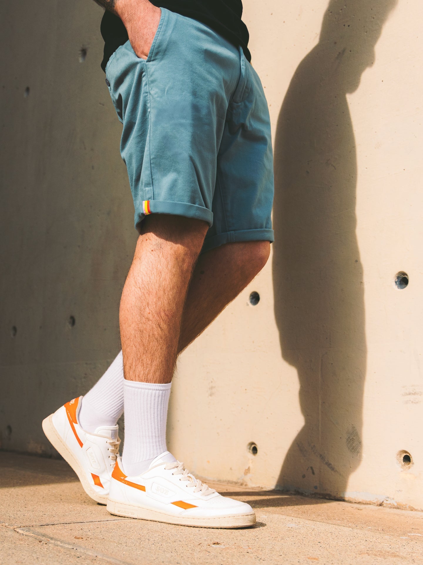 A person wearing La Machine Cycle Club Grand Tours - Rest Day Bermuda shorts in Antarctics stands against a beige wall, hands in pockets, legs crossed, paired with white socks and white sneakers with orange accents.