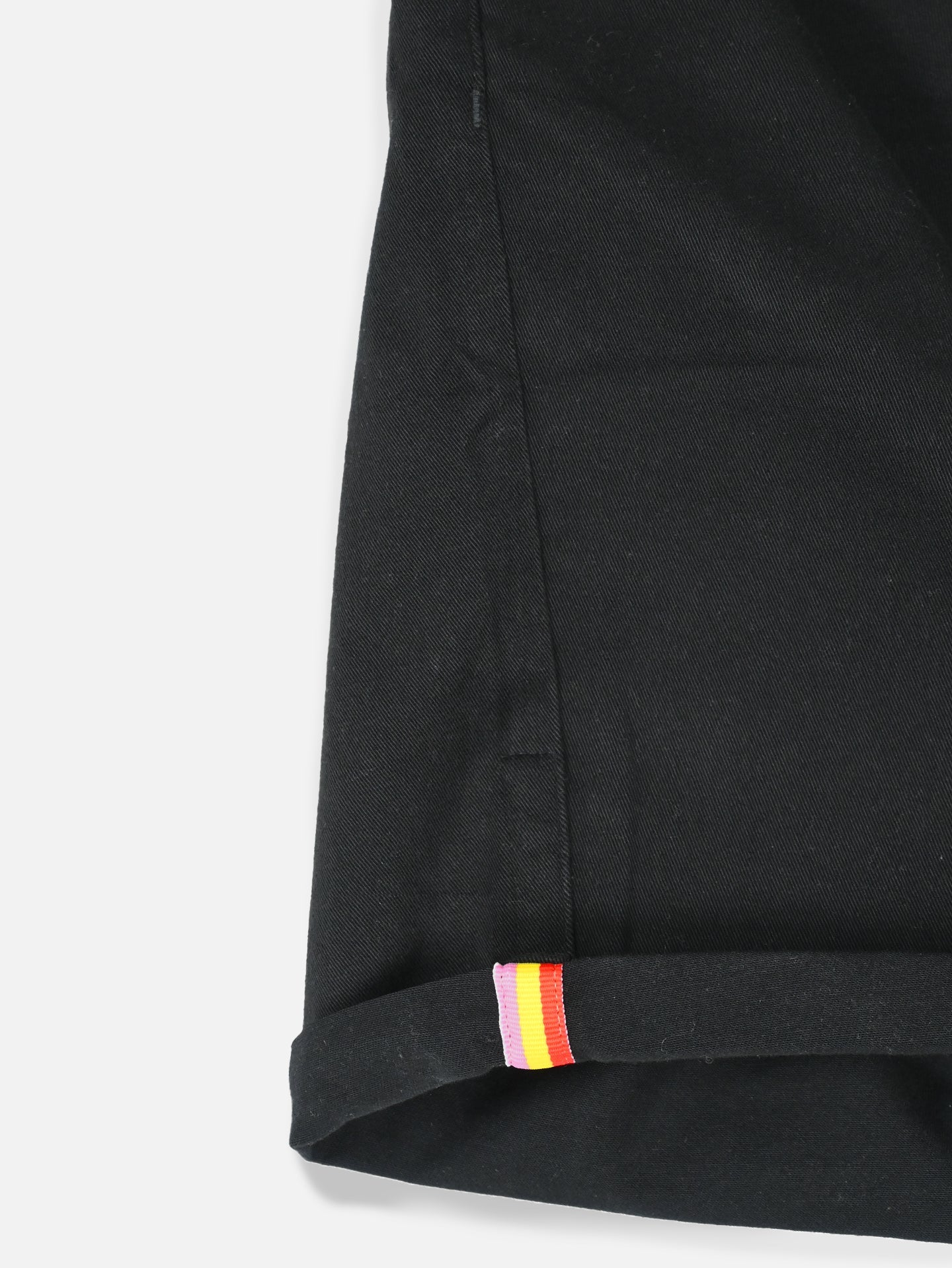 Close-up of the hem on La Machine Cycle Clubs Grand Tours - Rest Day Bermuda in black, showcasing a semi-tailored fit and a fabric tag with red, yellow, and pink stripes sewn into the cuff.
