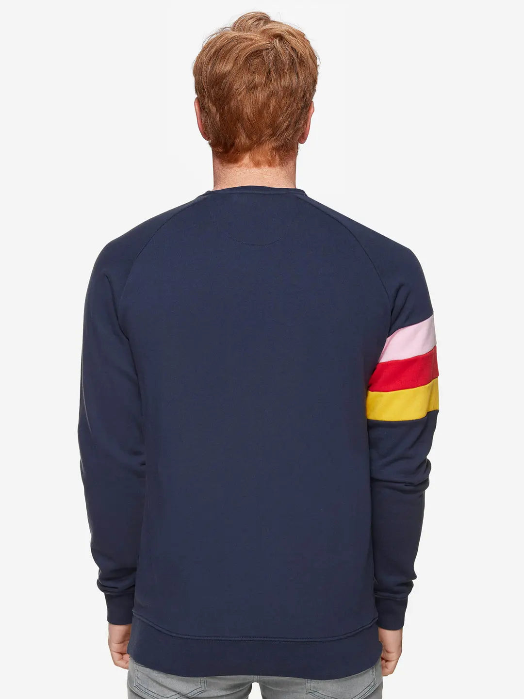 A person with light brown hair is seen from behind against a white background, wearing the La Machine Cycle Club Grand Tours Sweatshirt in navy blue, featuring pink, red, and yellow stripes on the right sleeve.