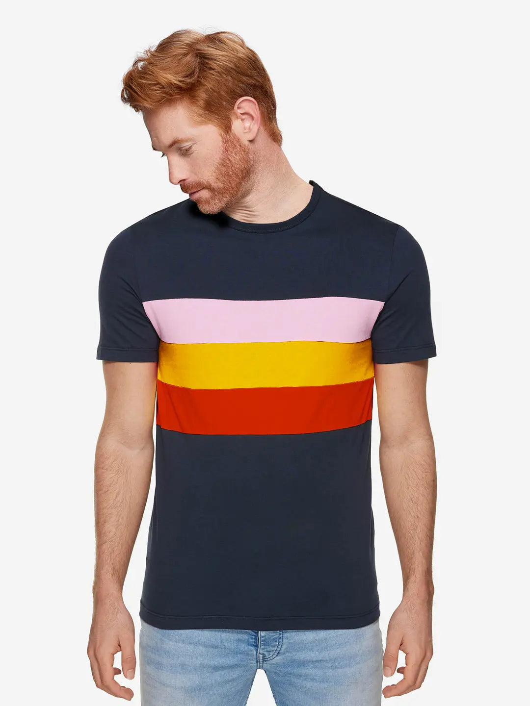 A light-skinned, red-haired man wears the La Machine Cycle Club Grand Tours T-shirt, featuring pink, yellow, and orange stripes, while standing against a plain white background and looking down to his left.