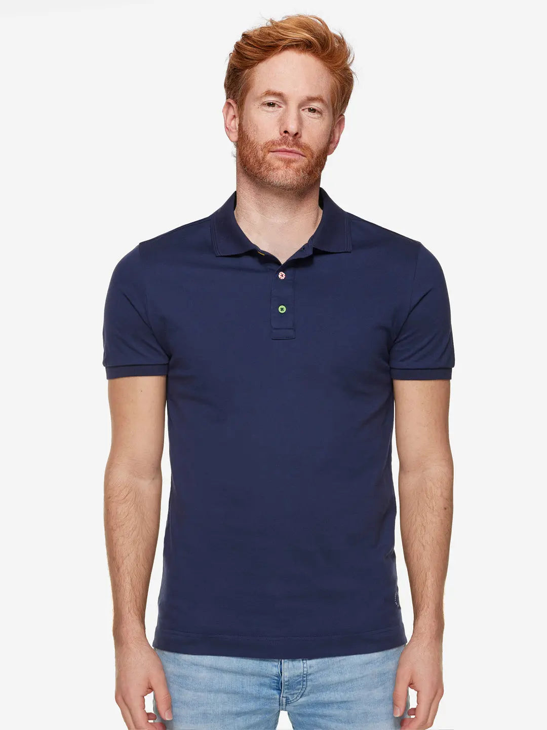 A light-skinned red-haired man wears the La Machine Cycle Club Grande Boucle Polo Shirt in navy with light blue jeans, standing against a plain white background and looking at the camera with a neutral expression.