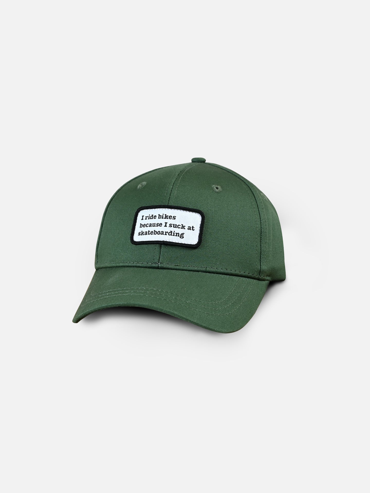 The La Machine Cycle Club Suck at Skateboarding - Casual Cap is a green adjustable bike cap with a front patch that reads, I ride bikes because I suck at skateboarding, shown on a plain white background.