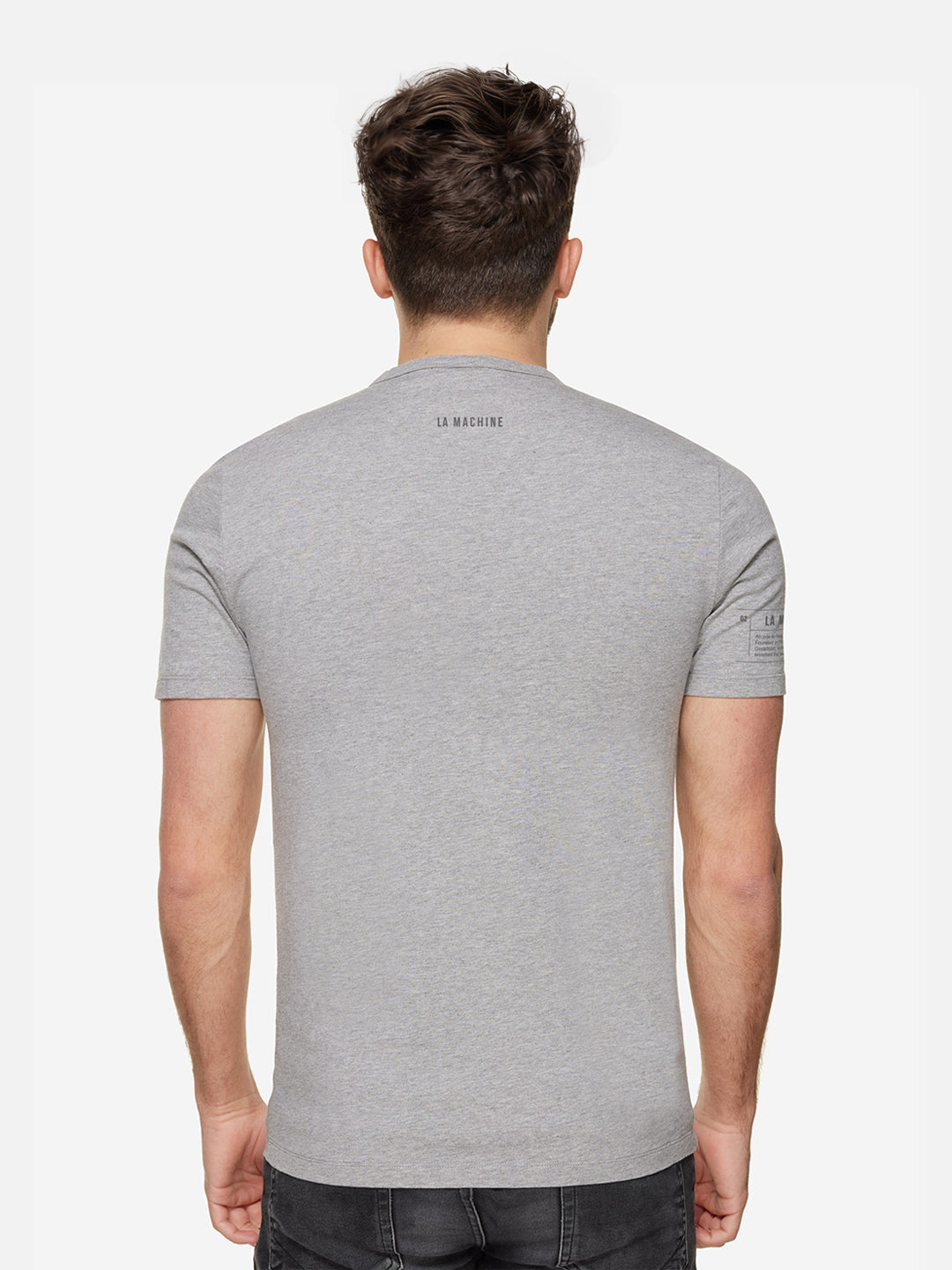 A man with short brown hair is facing away, wearing a fitted light grey Ijsboerke - The Remix T-shirt by La Machine Cycle Club, featuring LA MACHINE near the upper back. He pairs it with dark jeans against a plain white background.