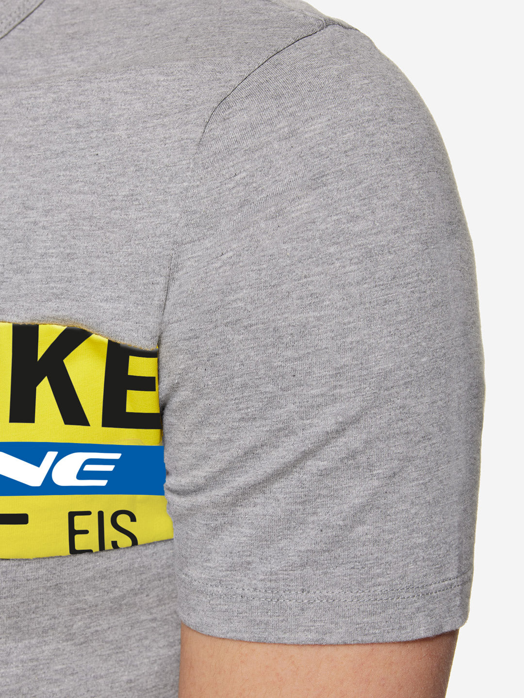 Close-up of a person’s upper arm and shoulder wearing La Machine Cycle Clubs Ijsboerke - The Remix T-shirt, a 100% cotton light grey unisex tee featuring a partial graphic with black, yellow, blue, and white text inspired by the Ijsboerke Cycling Team.