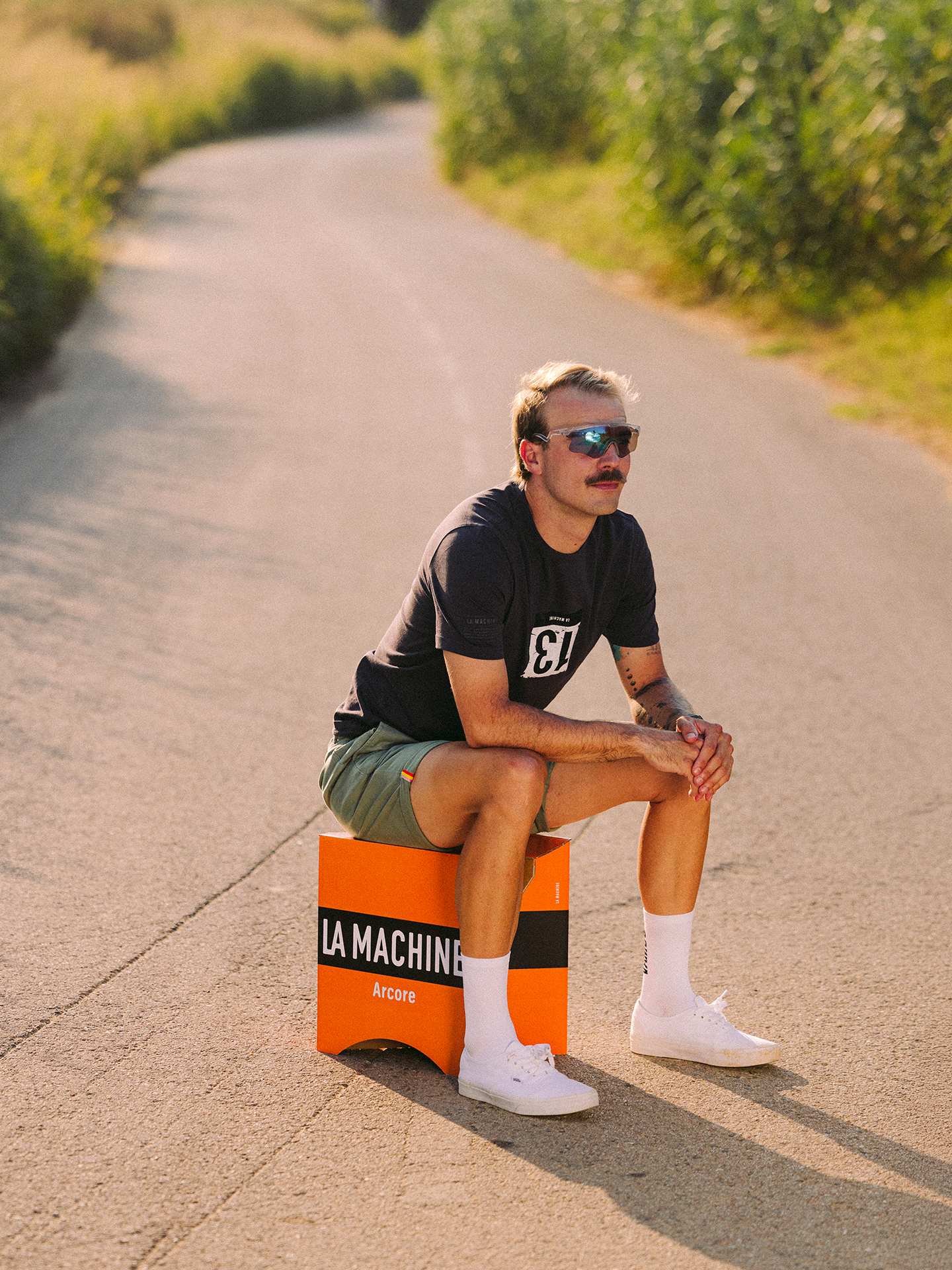 A man with blond hair, sunglasses, and a mustache sits on an orange Molteni 2-in-1 Stool & Sidetable by La Machine Cycle Club on a sunny, empty path. He wears a black t-shirt, green shorts, white socks, and white sneakers.