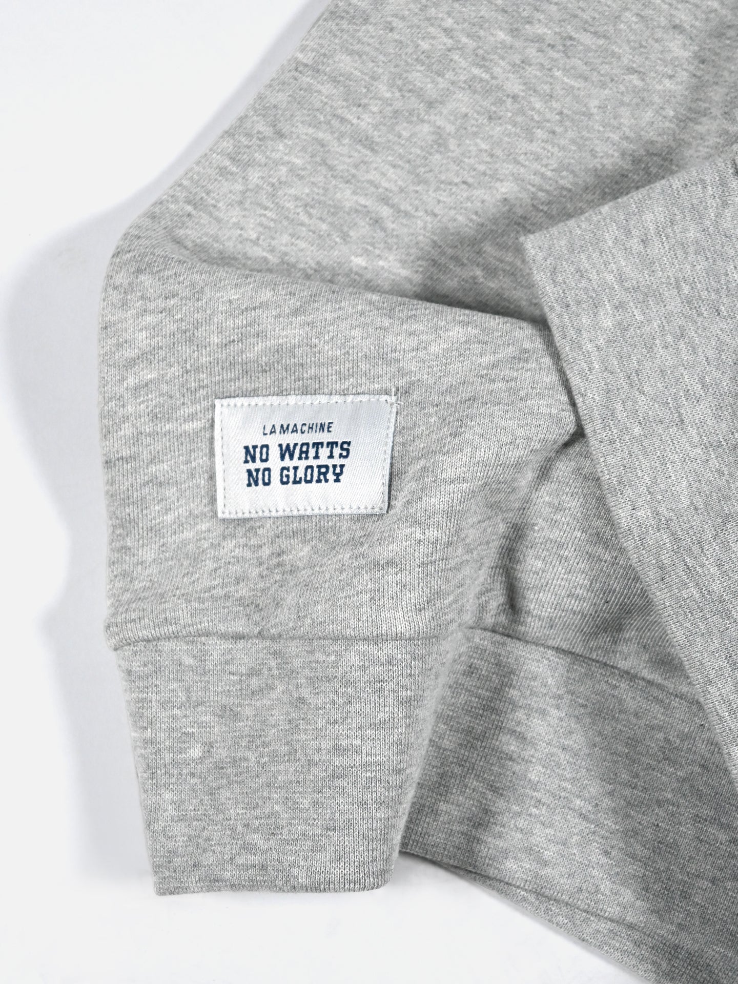 A close-up of the gray LMCC - University Hoodie by La Machine Cycle Club shows a soft, slightly folded sleeve with a white patch reading LA MACHINE NO WATTS NO GLORY in blue text.