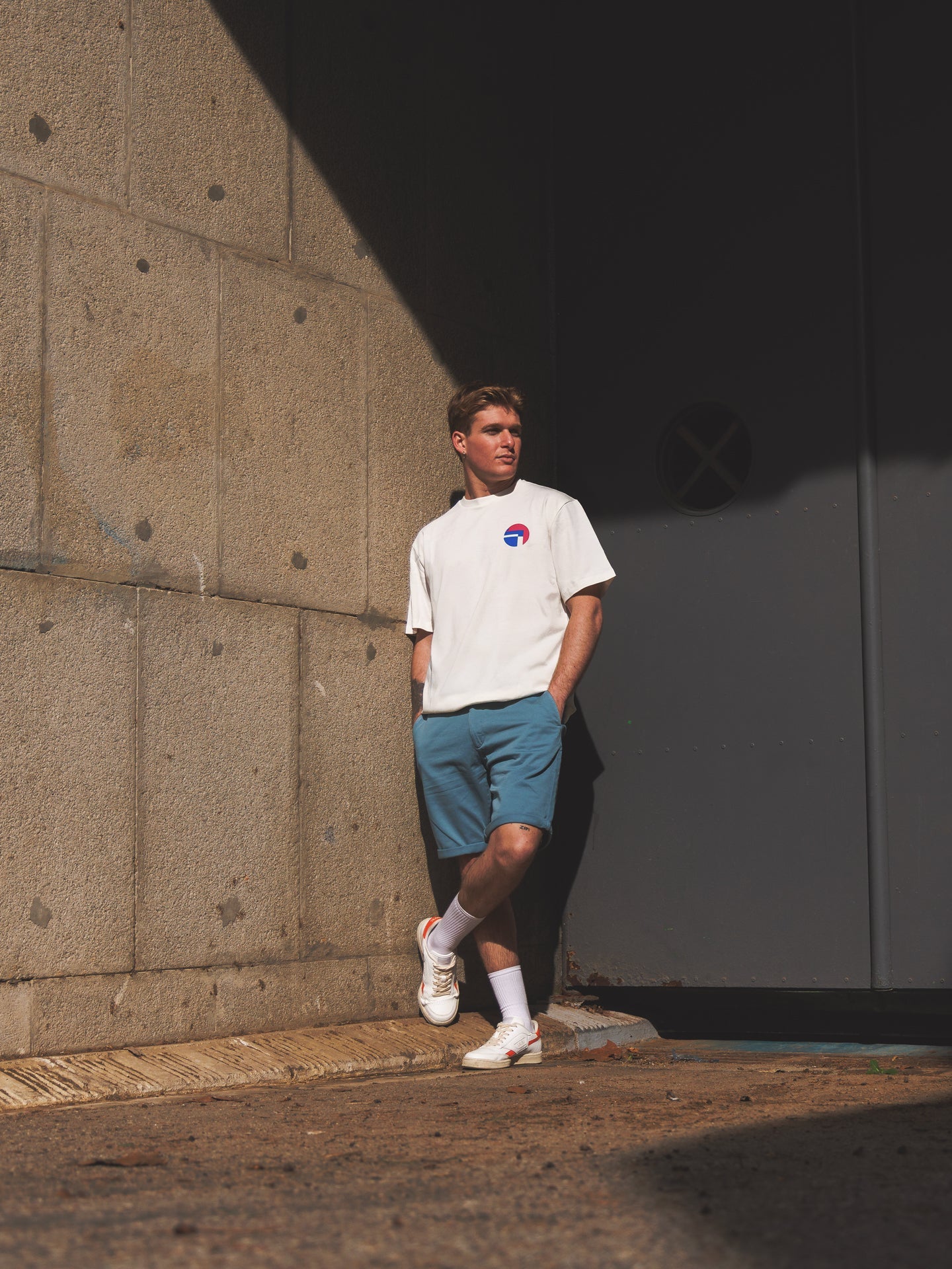 Wearing a Lampre Fondital 06 Loose Fit T-shirt by La Machine Cycle Club, a man leans against a concrete wall in partial sunlight, hands in pockets, looking thoughtfully into the distance.