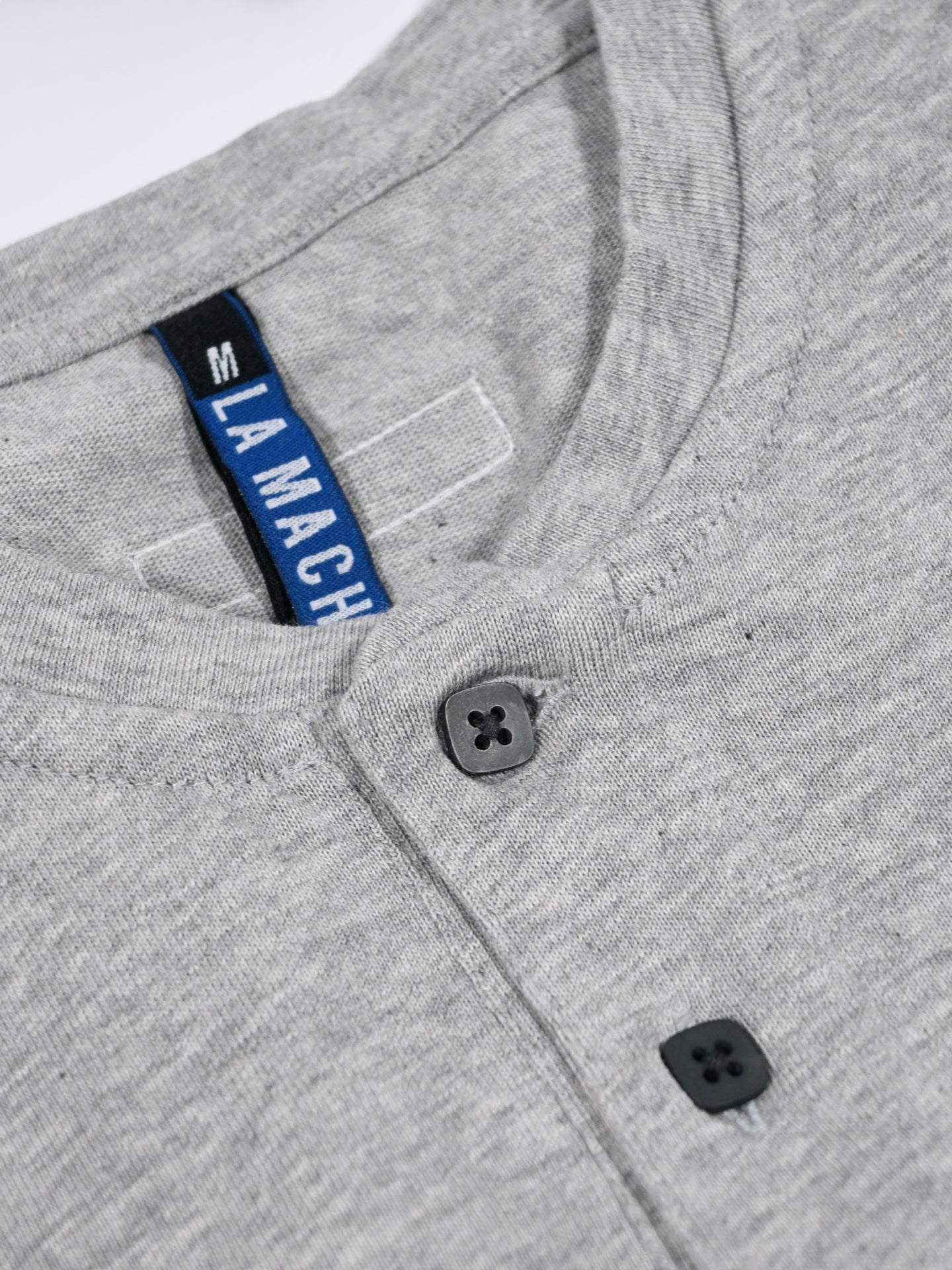 A close-up of the neckline of the Le Pavé - Henley in Light Cobble Grey by La Machine Cycle Club, featuring two black buttons and a tag with La Machine in white letters on a blue and black background.