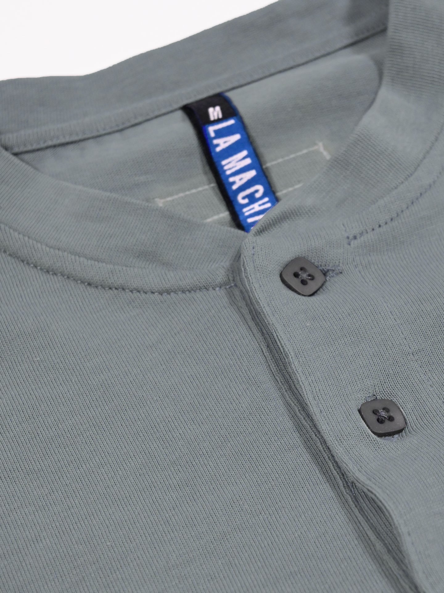 A close-up of the Le Pavé Henley in Roubaix Blue from La Machine Cycle Club shows two neckline buttons and a visible blue and black LA MACHINE label.