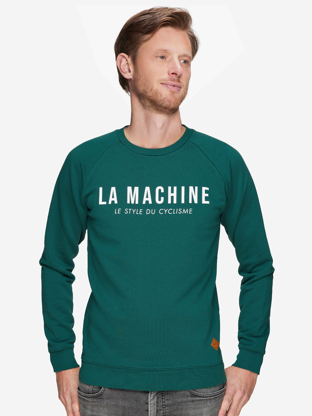 A man wearing a teal La Machine Cycle Club La Machine Logo - Sweatshirt with LE STYLE DU CYCLISME on the front stands against a white background, looking to the side with a slight smile.