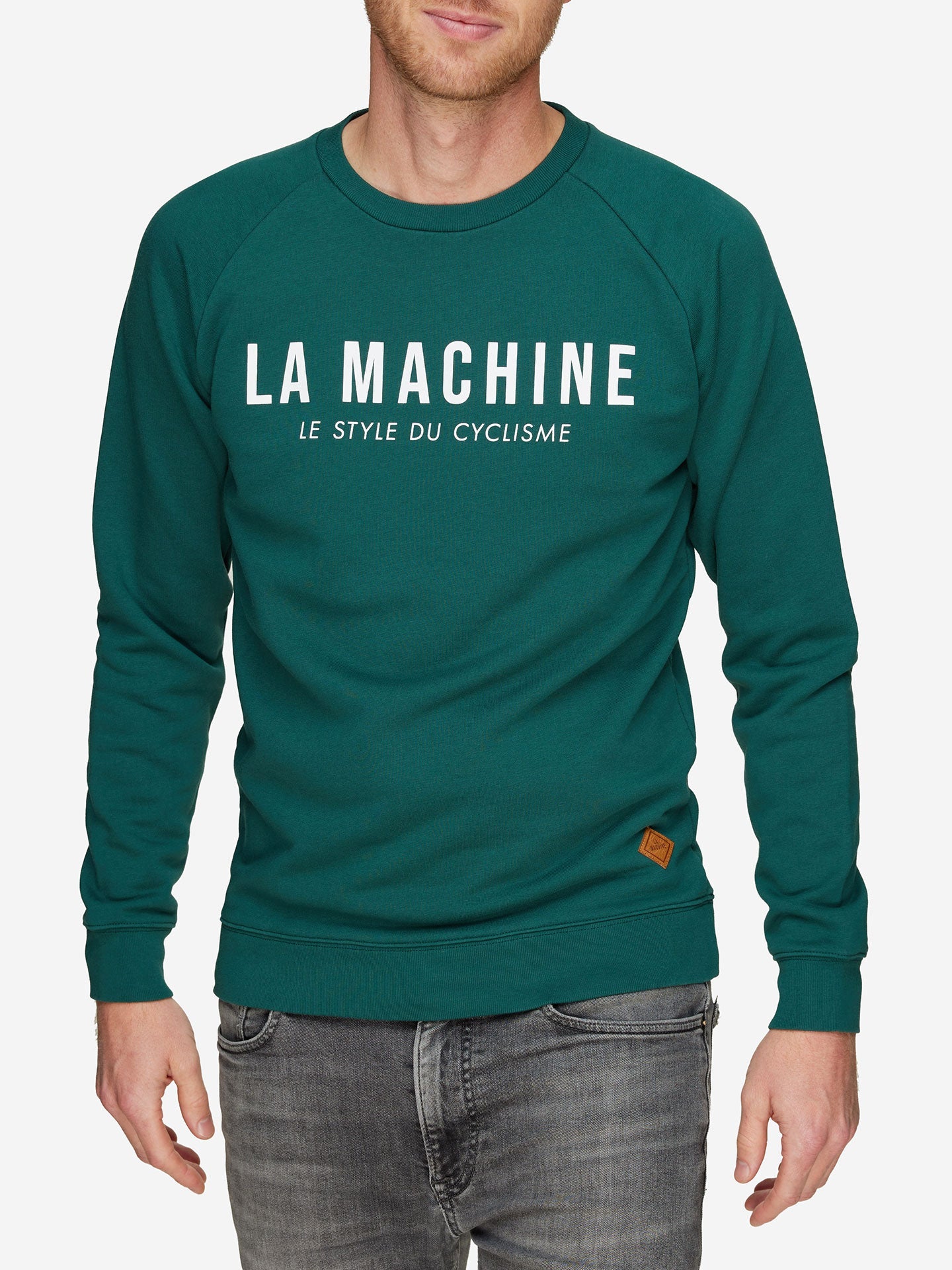 A man wears a teal La Machine Logo - Sweatshirt from La Machine Cycle Club, featuring LA MACHINE LE STYLE DU CYCLISME in white on the chest, paired with grey jeans. The background is plain white.