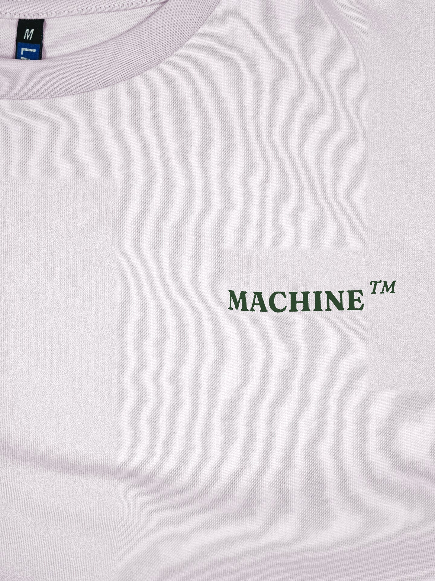 A close-up of the La Machine Cycle Club Made in Mons-en-Pévèle - Loose Fit - T-shirt shows MACHINE™ in dark green on the left chest and an M size tag visible inside the collar.