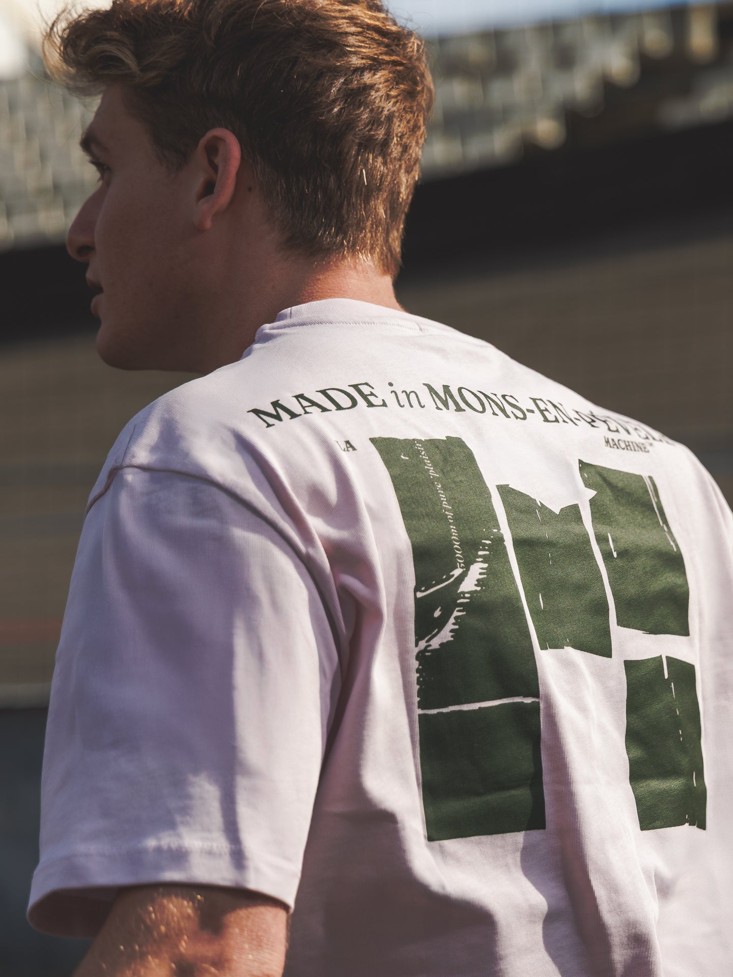 A person with short, light brown hair wears the La Machine Cycle Club Made in Mons-en-Pévèle loose fit T-shirt—light pink with green graphics—great cycling wear for a sunny ride near Mons-en-Pévèle.