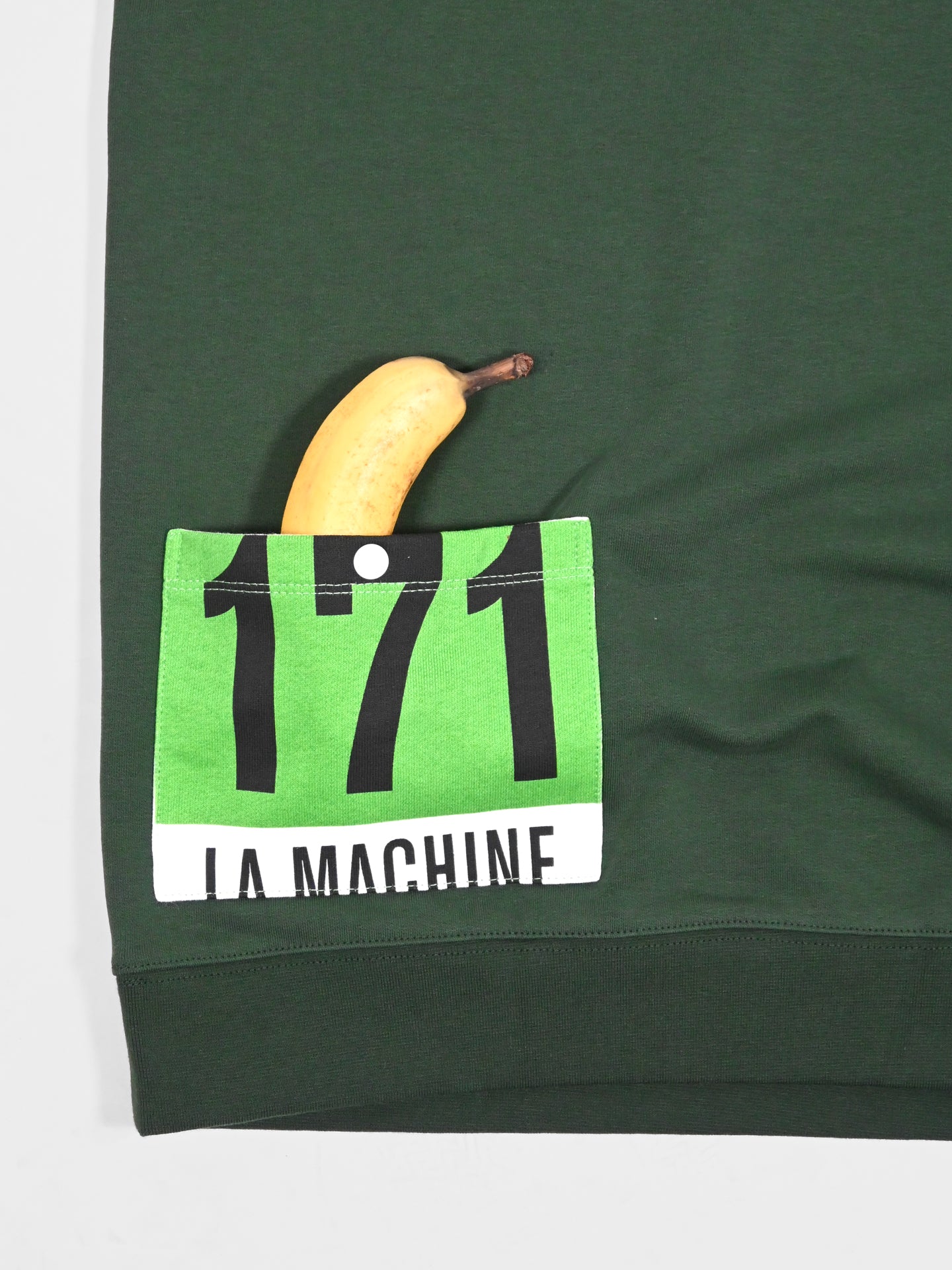 A yellow banana peeks from a green race bib #171, pinned to the pocket of the Maillot Vert - Sweatshirt by La Machine Cycle Club. The bib reads LA MACHINE. Made in EU from 100% cotton.