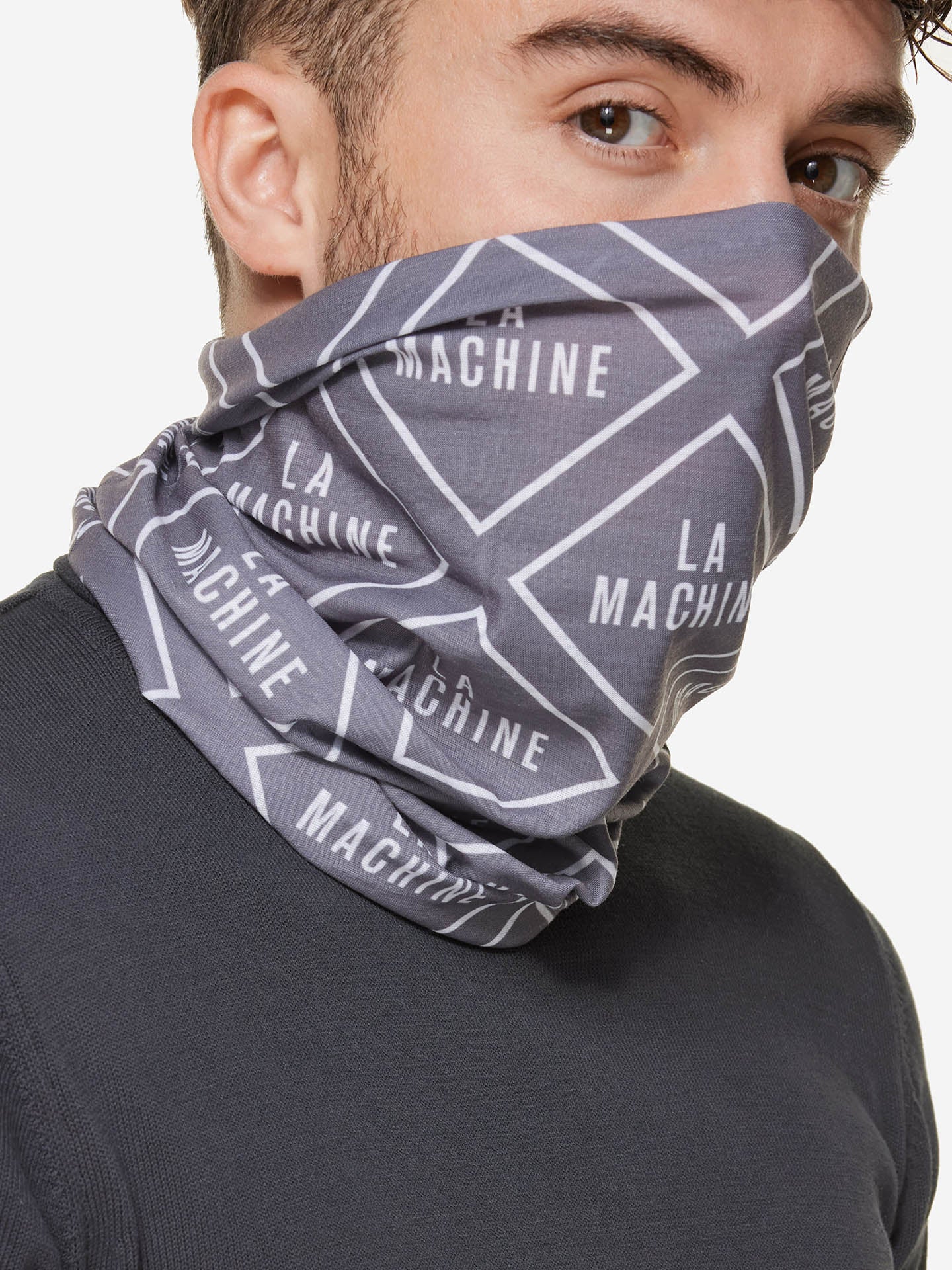 A man with short brown hair, dressed in a dark grey shirt, wears the La Machine Cycle Club Neck Warmer – Grey, featuring white geometric patterns and repeated “LA MACHINE” branding, covering his neck and lower face.