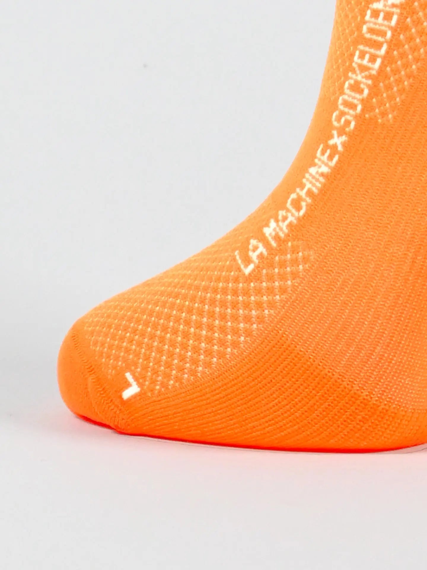 Close-up of the No Watts No Glory - Cycling Socks in orange by La Machine Cycle Club, featuring white “LA MACHINE X SOCKELER” text and a small “L” near the toes. Made in Italy with a seamless toe, shown against a plain light background.