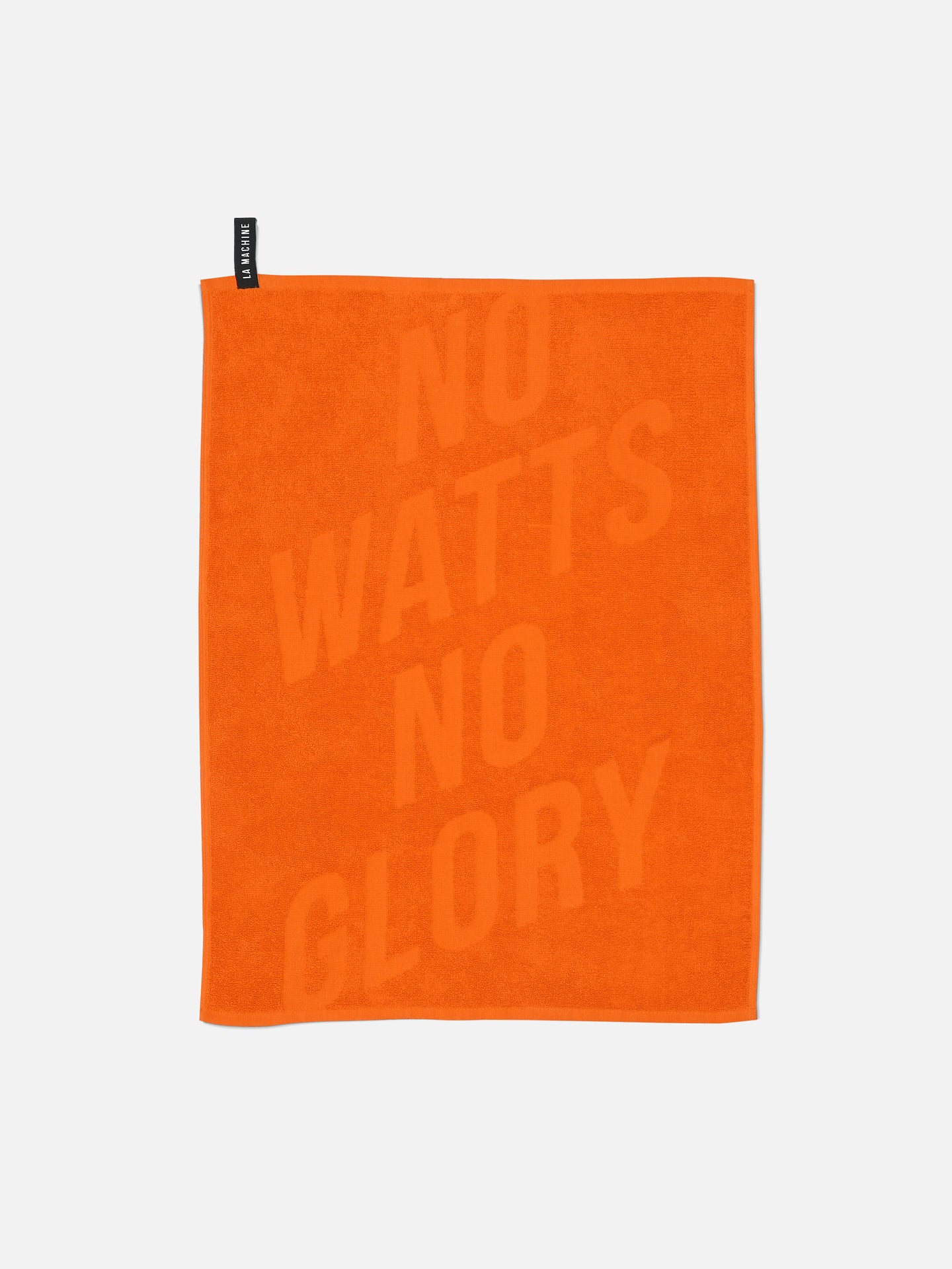 The La Machine Cycle Club No Watts No Glory Towel (60x80 cm) is orange, made of 100% cotton, and features the slogan printed diagonally. A black tag accents the upper left edge. The background is plain white.