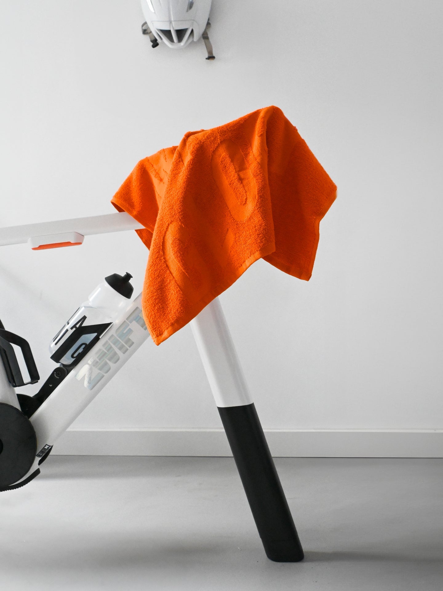 A No Watts No Glory Towel by La Machine Cycle Club, 60x80 cm, in bright orange drapes over a stationary bike’s handlebars; a white water bottle and helmet hang in the background, ready for your next virtual ride.