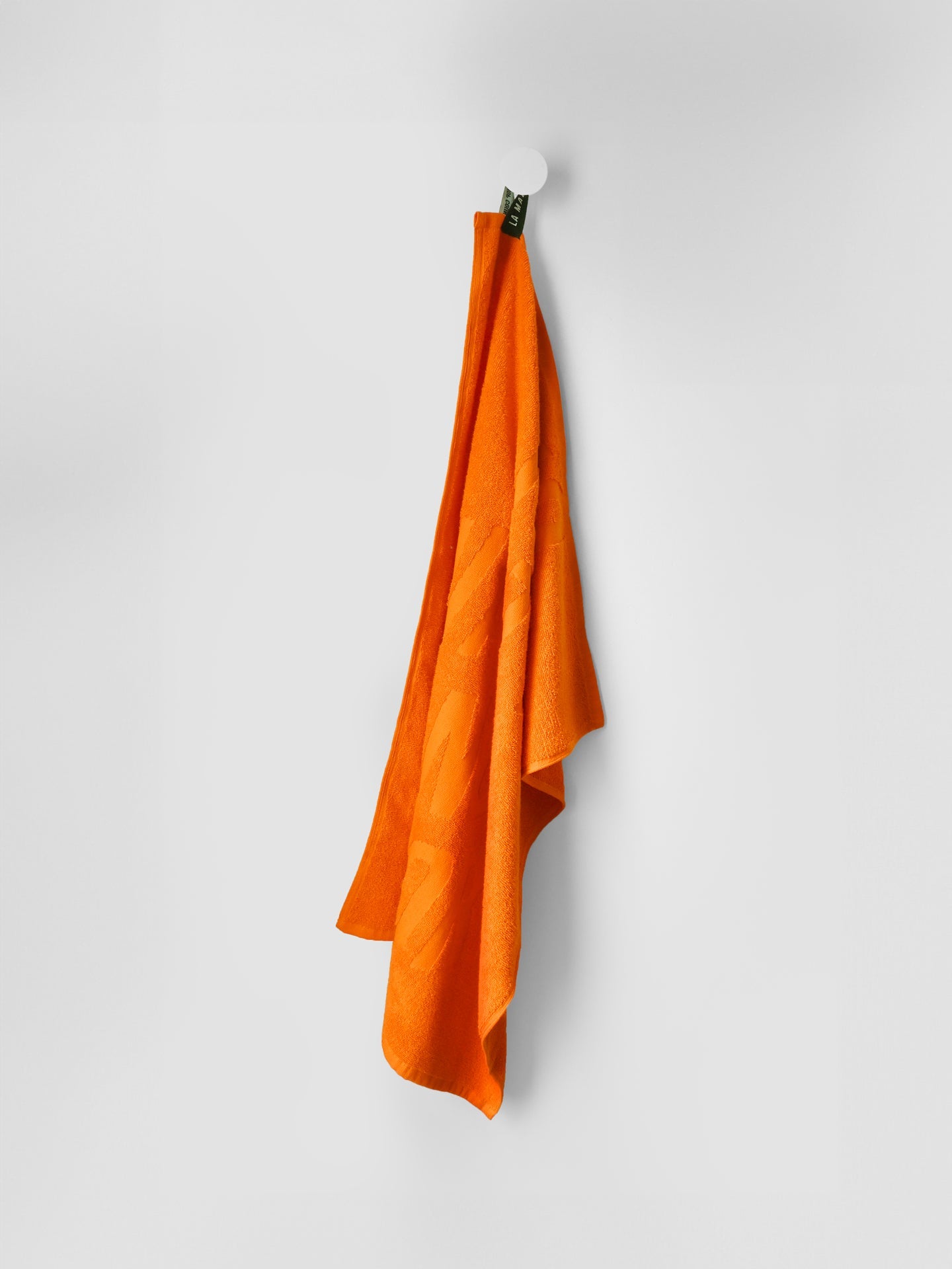 A No Watts No Glory towel by La Machine Cycle Club (60x80 cm, 100% cotton) hangs neatly on a white round hook against a plain white wall.