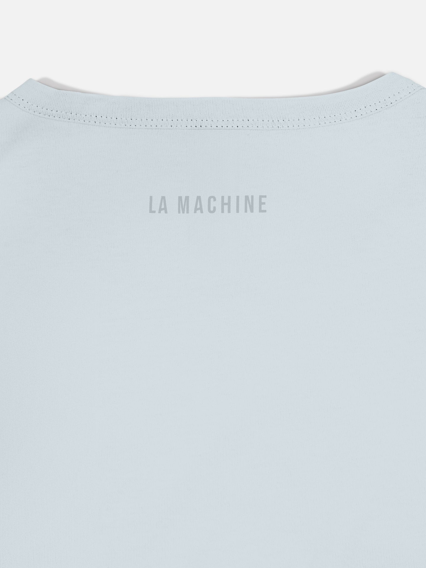 Close-up of the upper back of the Pedaleur de Charme - Embro T-shirt by La Machine Cycle Club, a light blue, 100% cotton unisex tee featuring LA MACHINE in small gray uppercase letters near the neckline.
