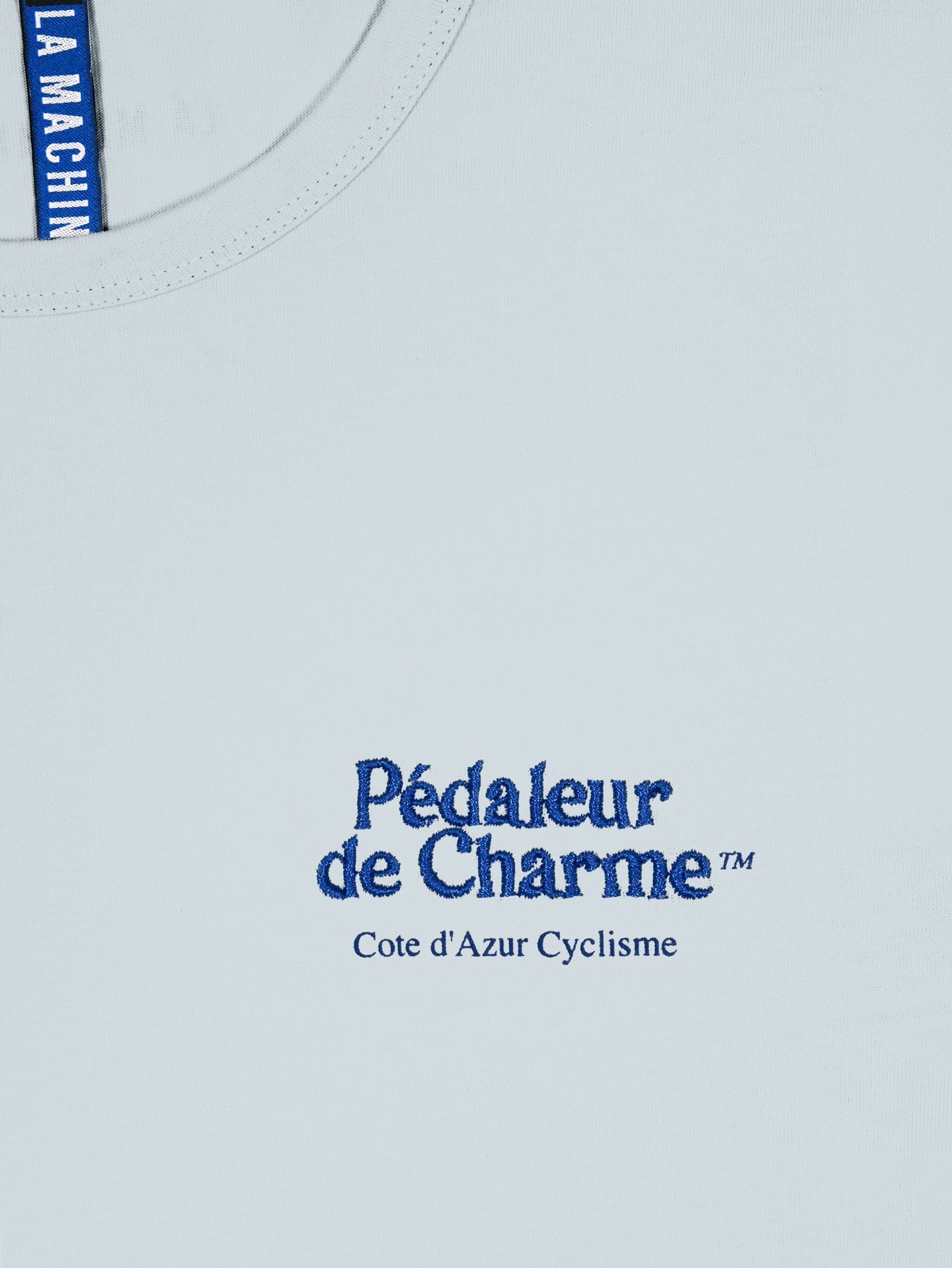 A close-up of the Pedaleur de Charme - Embro - T-shirt by La Machine Cycle Club, featuring blue Pédaleur de Charme™ and Cote dAzur Cyclisme text on the chest, a blue LA MACHINE tag at the neckline, and made from 100% cotton.
