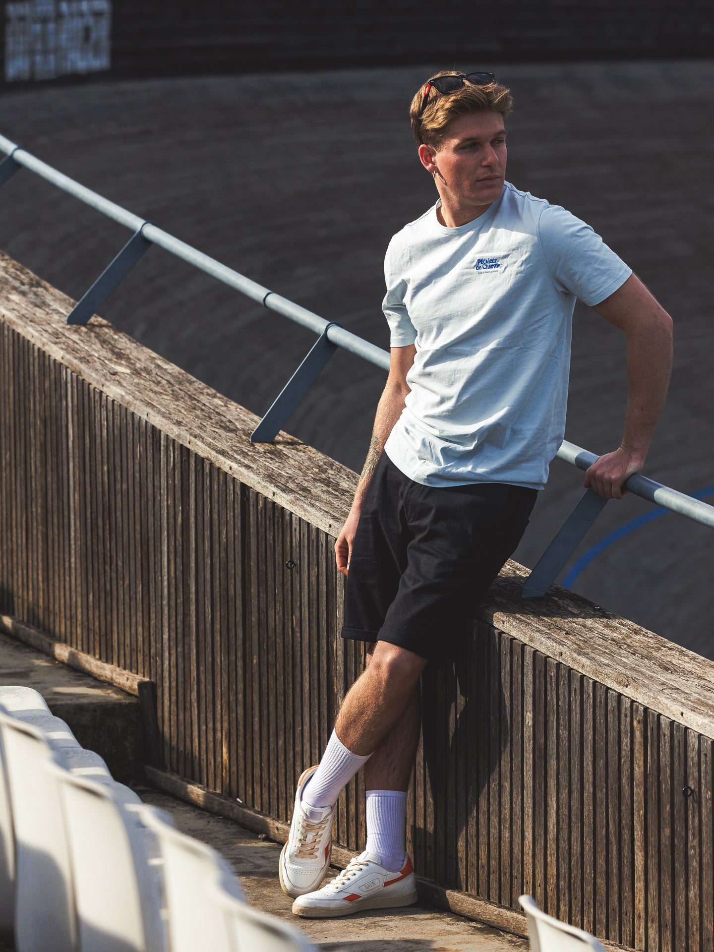A man in a La Machine Cycle Club Pedaleur de Charme - Embro T-shirt leans against a wooden railing outdoors, wearing black shorts and white sneakers, with empty stadium seats and a curved track in the background.