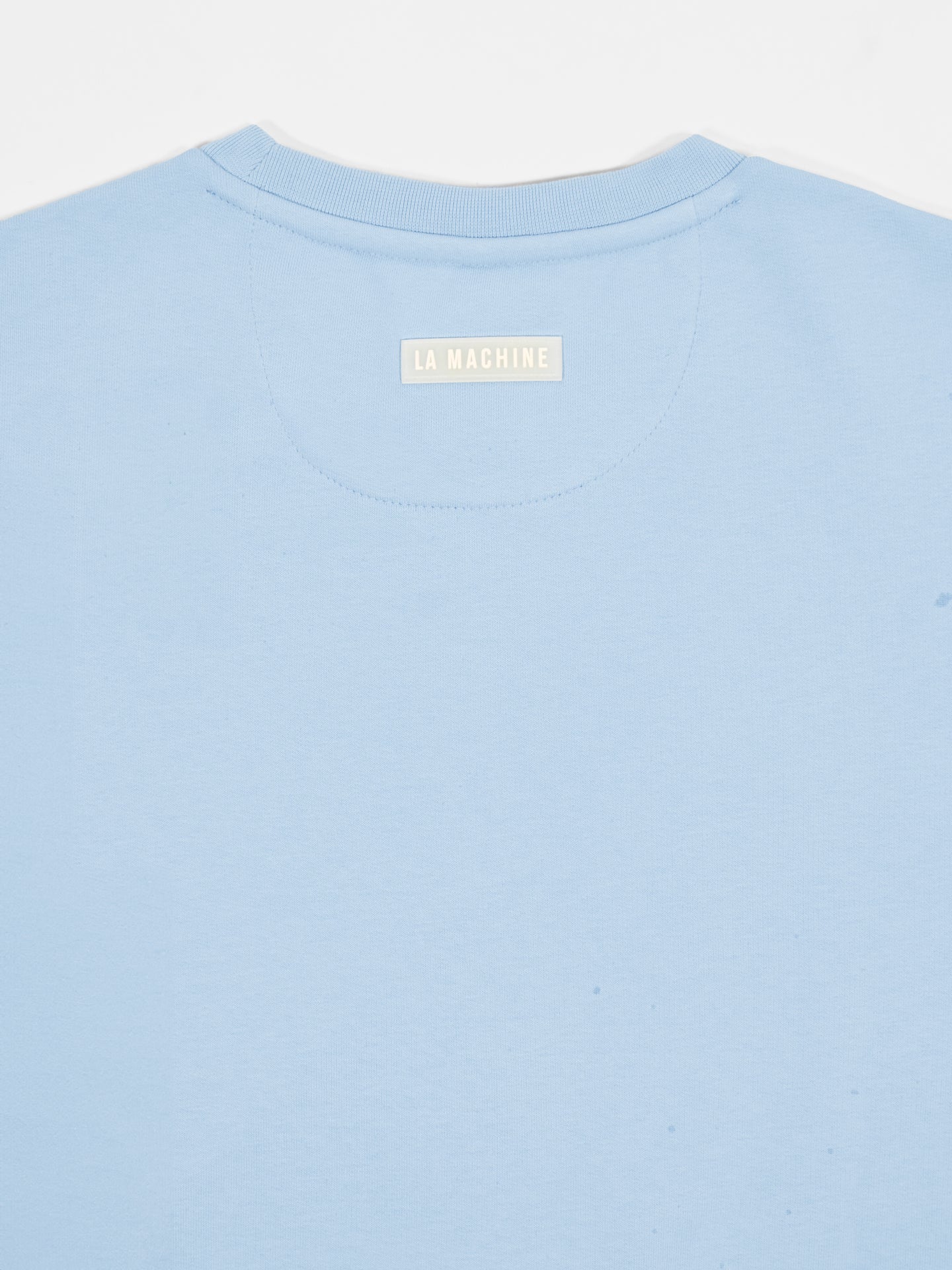 A close-up of the back neckline of the Pedaleur de Charme - Sweatshirt in light blue by La Machine Cycle Club, featuring a beige LA MACHINE label stitched below the collar, inspired by Giro dItalia winner Hugo Koblet.