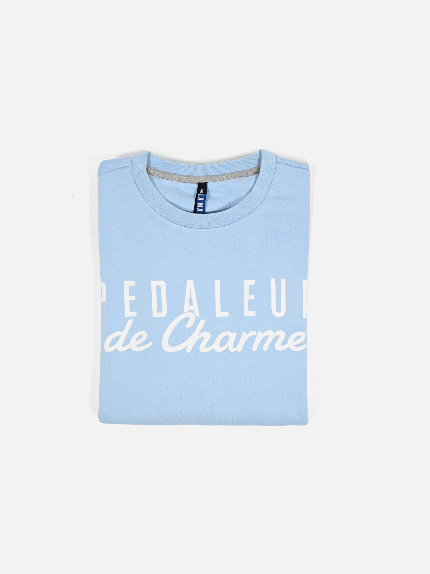 A light blue “Pedaleur de Charme” sweatshirt by La Machine Cycle Club, inspired by Giro d’Italia winner Hugo Koblet, is shown folded with white print on a plain white background.