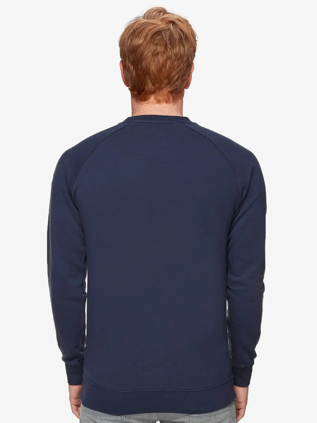 A person with short, light brown hair is seen from behind against a white background, wearing La Machine Cycle Club’s Pedaleur de Charme Sweatshirt in navy and light gray jeans—a look inspired by Giro d’Italia winner Hugo Koblet.