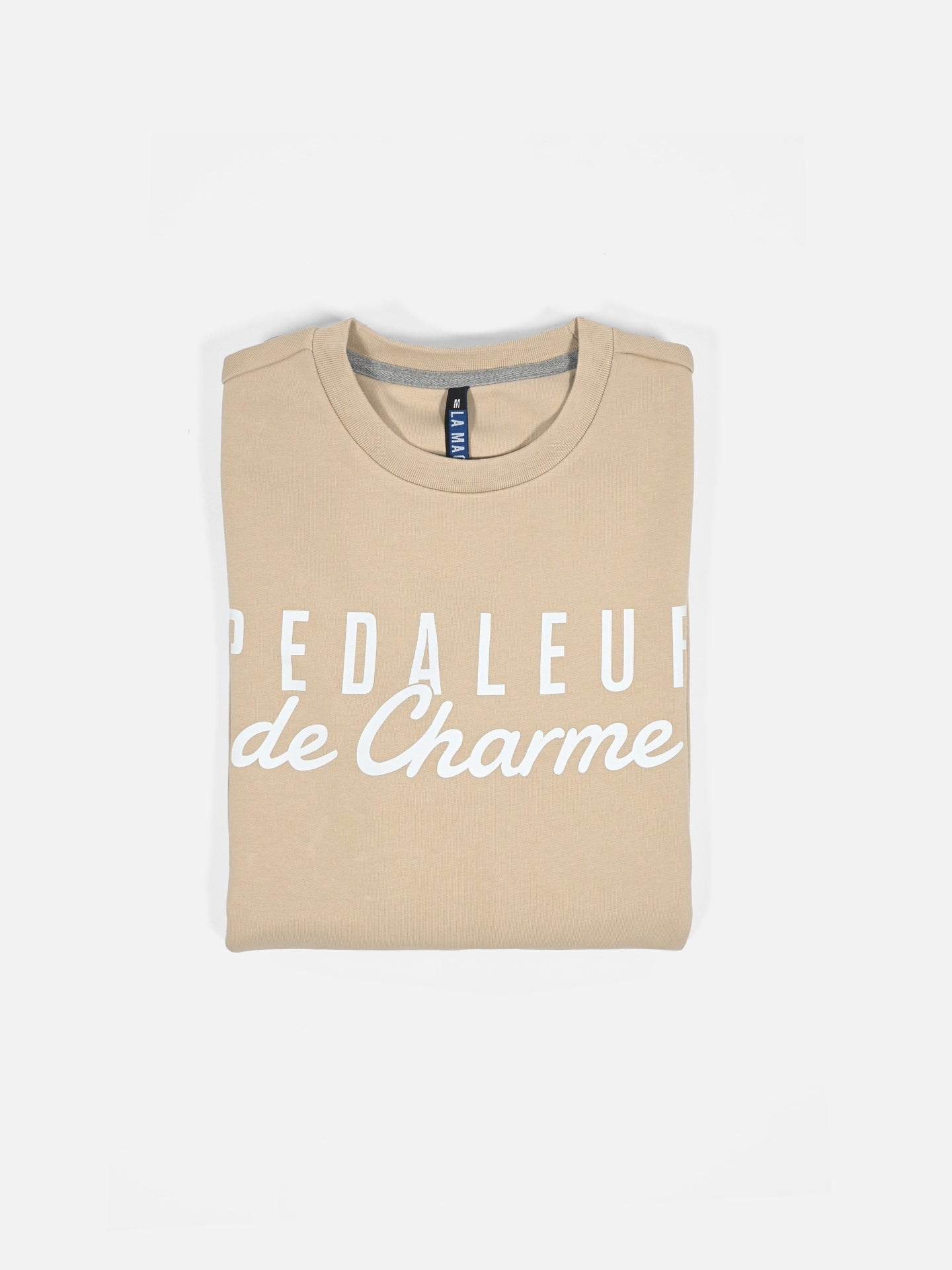 A sand-colored mens sweatshirt from La Machine Cycle Club, neatly folded, featuring Pedaleur de Charme in white—a stylish tribute to Giro d’Italia apparel and the classic Hugo Koblet look.