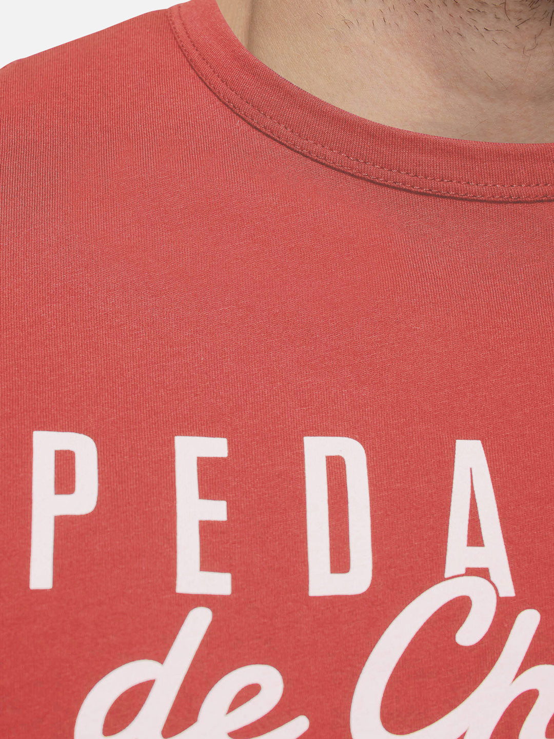 Close-up of a person wearing the La Machine Cycle Club Pedaleur de Charme T-shirt in coral, featuring partially visible white text. The image shows the top of their chest, neck, and jawline.