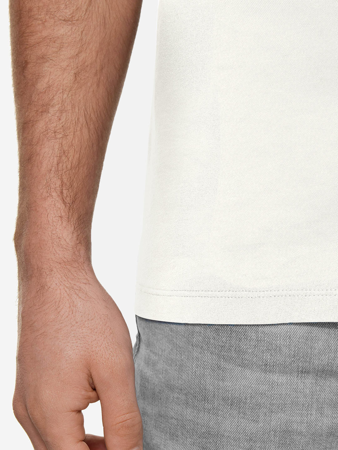 A close-up of a person’s left hand and forearm beside their torso, wearing the La Machine Cycle Club Pedaleur de Charme T-shirt in Off White and light gray pants, highlights the premium fabric texture and natural arm hair.