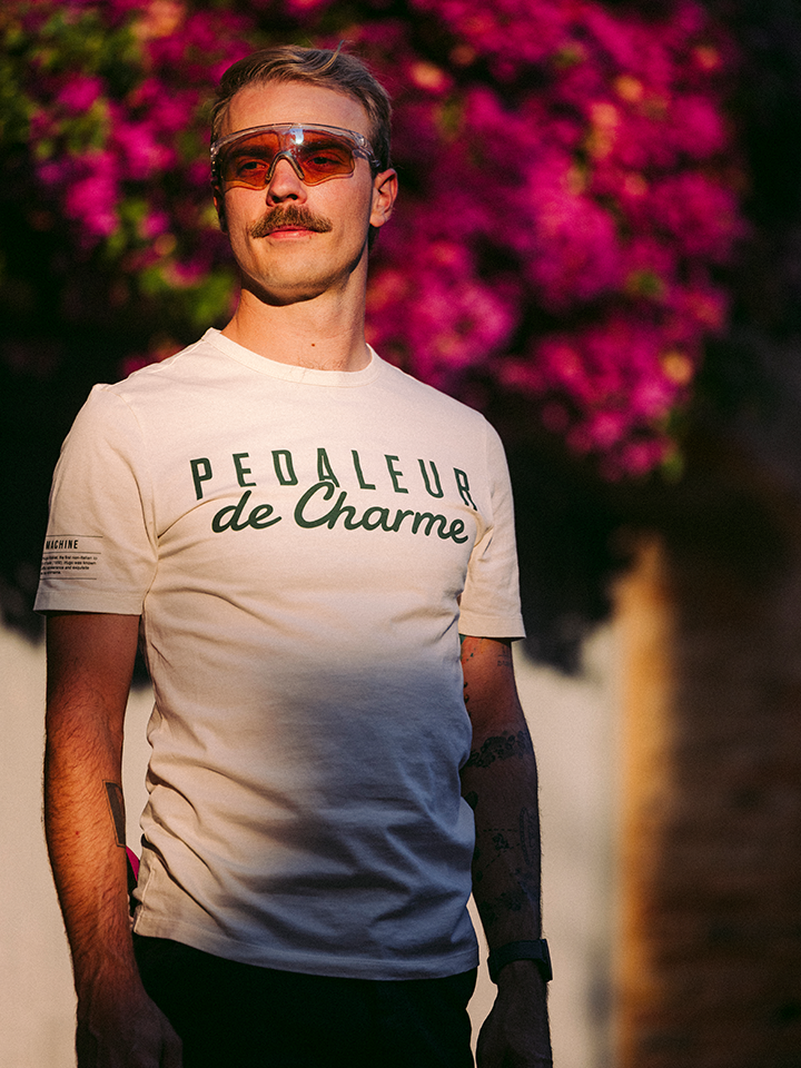 A man with a mustache wears orange sunglasses and an off-white Pedaleur de Charme t-shirt by La Machine Cycle Club, standing in sunlight with vibrant purple flowers blurred in the background.