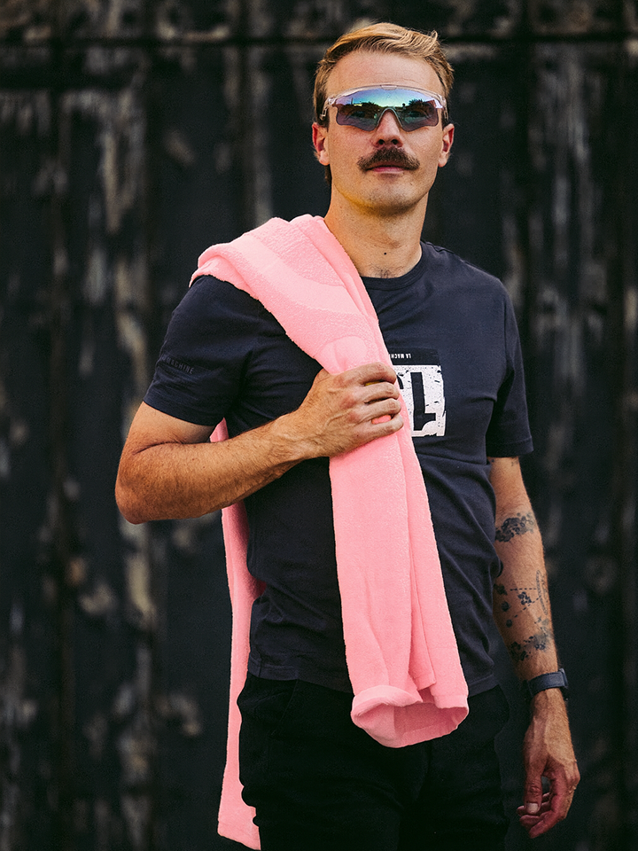 A man with blond hair and a mustache, wearing sunglasses and a black t-shirt, holds the La Machine Cycle Club Il Giro della Spiaggia Beach Towel over his shoulder while standing before a weathered dark wooden background.
