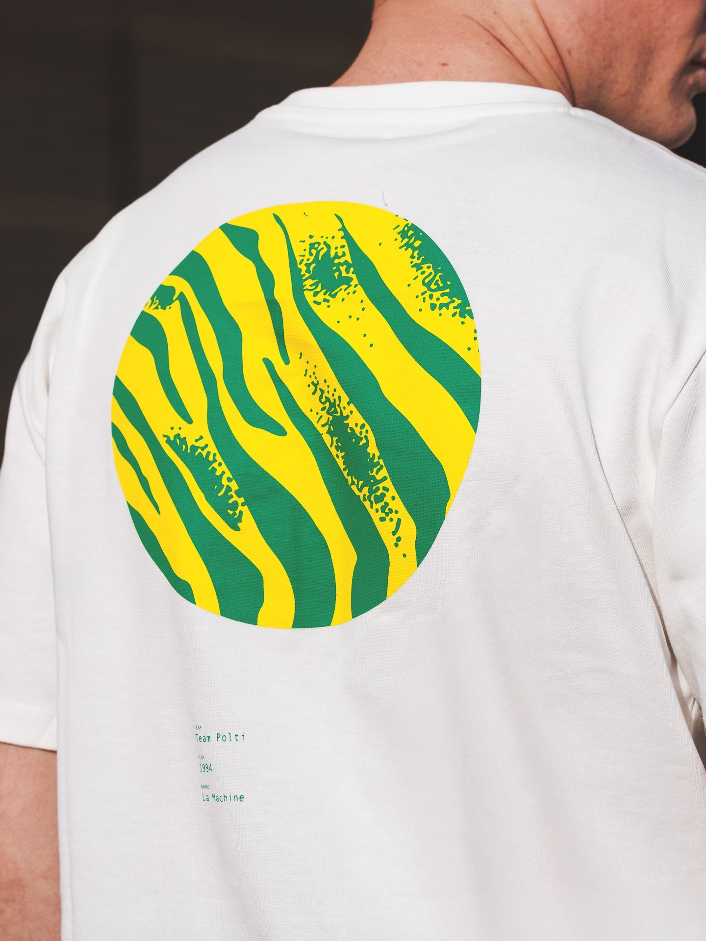 A person seen from the back and side wears the La Machine Cycle Club Team Polti 94 Loose Fit T-shirt, featuring a large circular green and yellow wavy tiger-stripe pattern on the back.
