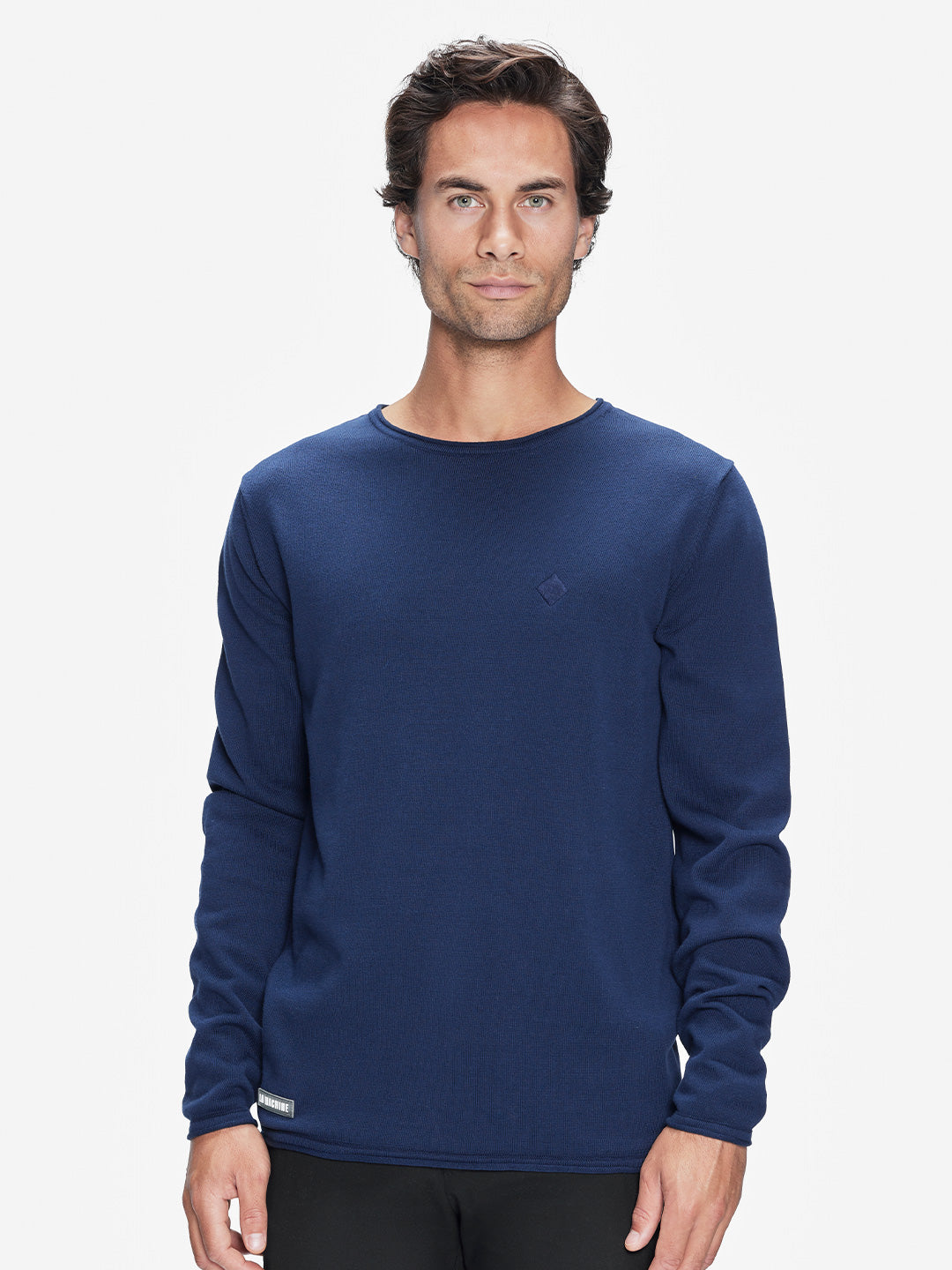 A man with wavy brown hair is wearing the La Machine Cycle Club Premium Knitted Pullover in navy blue with black pants, standing against a plain white background and looking directly at the camera with a neutral expression.