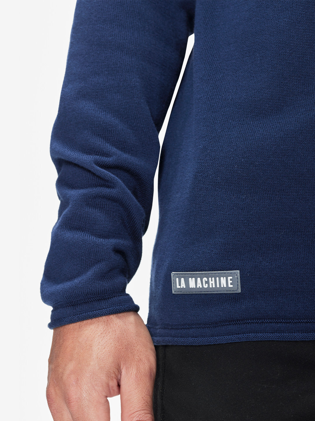 A close-up of a person’s arm and hand wearing the La Machine Cycle Club Premium Knitted Pullover in navy blue, featuring a “LA MACHINE” label near the hem, paired with black pants.
