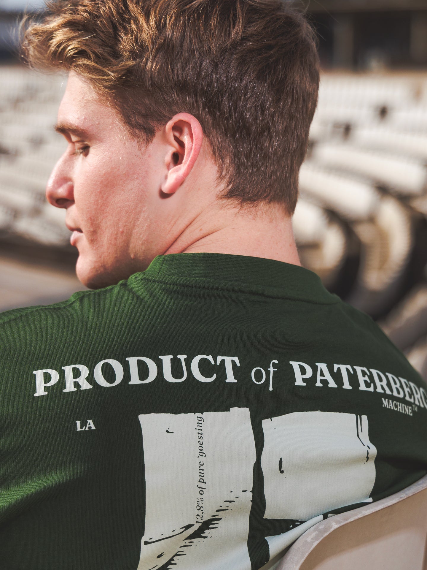 A man with light brown hair sits on a white stadium seat, showing the back of his dark green Product of Paterberg - Loose Fit T-shirt by La Machine Cycle Club, with bold white text—a tribute to legendary Flanders climbs.