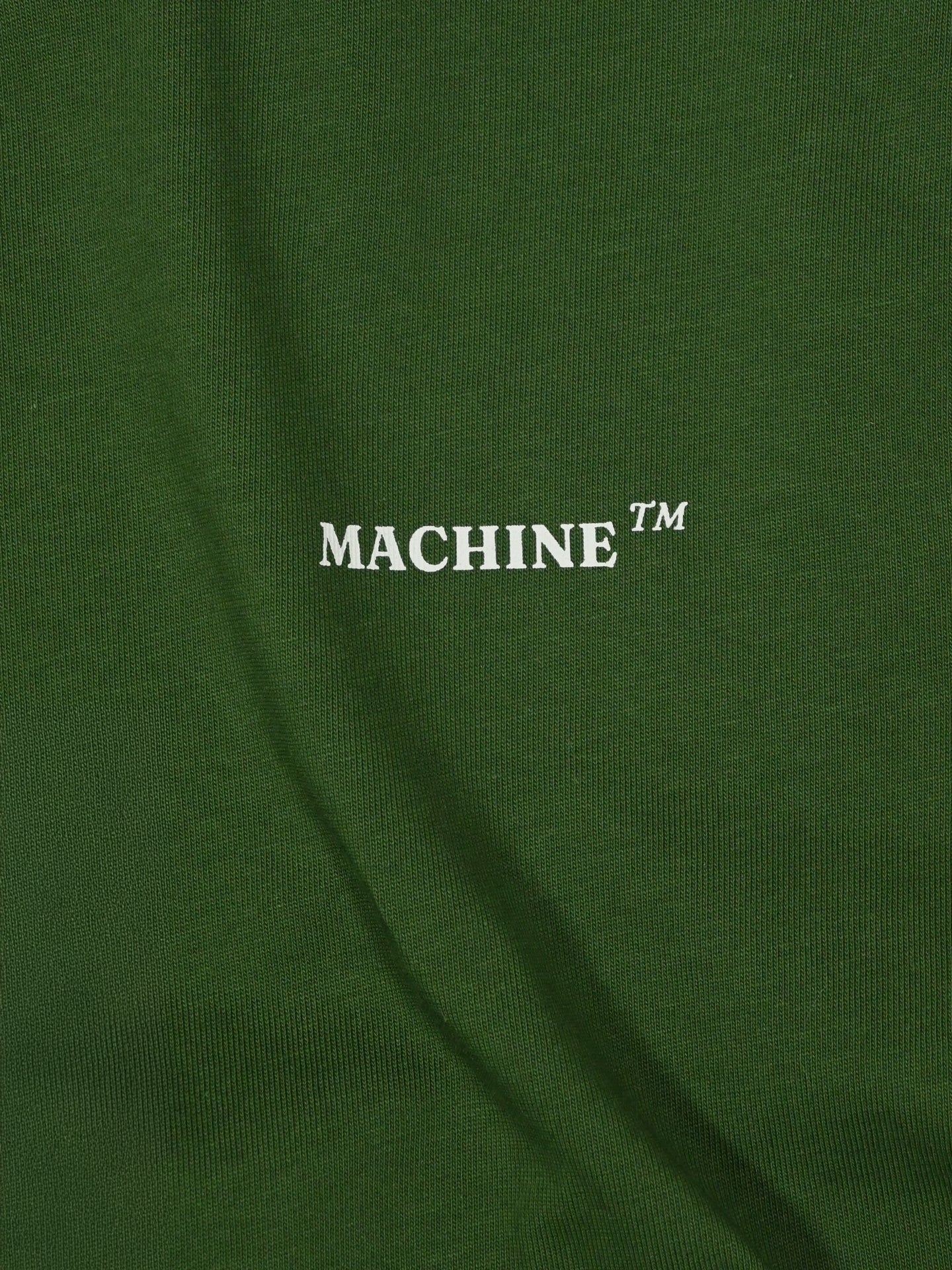 A close-up of dark green heavy cotton from the La Machine Cycle Club Product of Paterberg - Loose Fit - T-shirt shows MACHINE™ in bold white text, subtle folds, and a smooth finish inspired by legendary Flanders climbs.