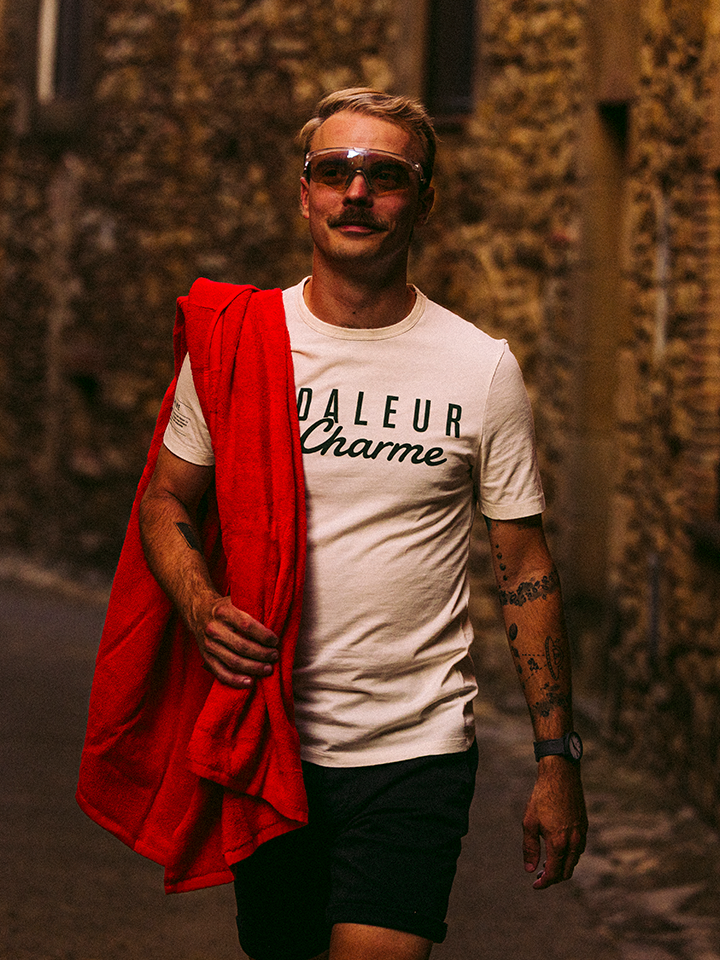 A mustachioed man in sunglasses, a “DALEUR Charme” tee, and black shorts walks a narrow street. He carries a red jacket and La Machine Cycle Club’s La Vuelta a la Playa beach towel over his shoulder, tattoos on his left arm.