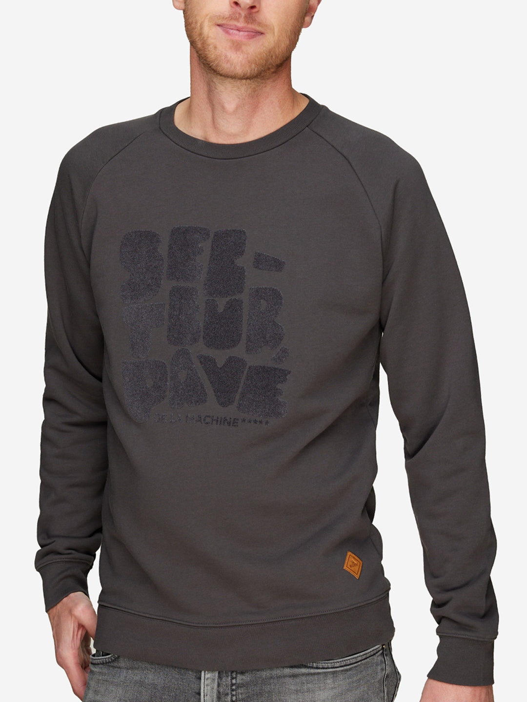 A man wears a dark gray Secteur Pavé - Sweatshirt by La Machine Cycle Club with faded Paris-Roubaix graphics and a brown patch near the left hem, paired with gray jeans. The background is white.