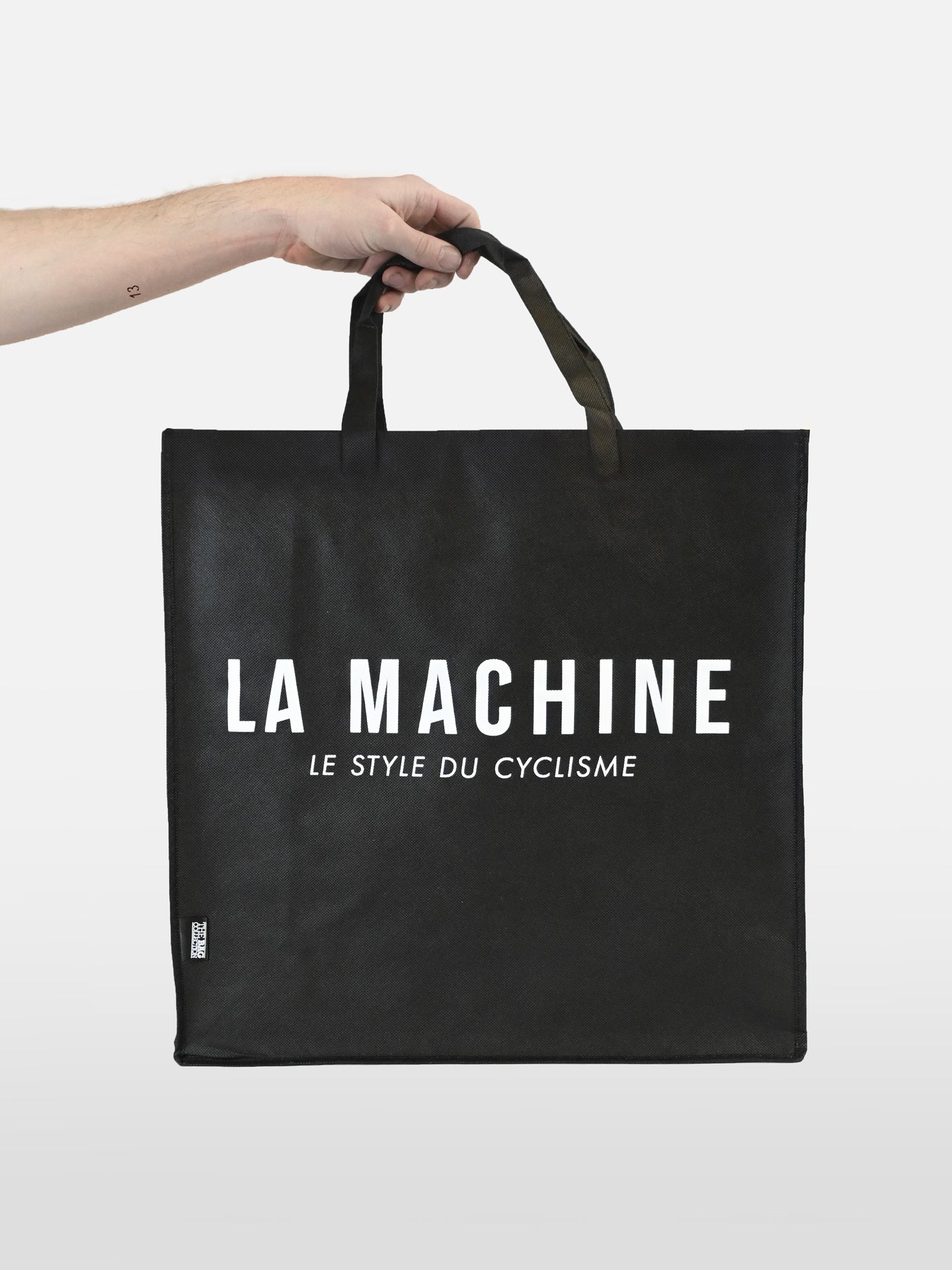 A hand holds the La Machine - Shopper by La Machine Cycle Club, a black non-woven bag with “LA MACHINE LE STYLE DU CYCLISME” in bold white letters on the front. The plain white background highlights this stylish cycling accessory.