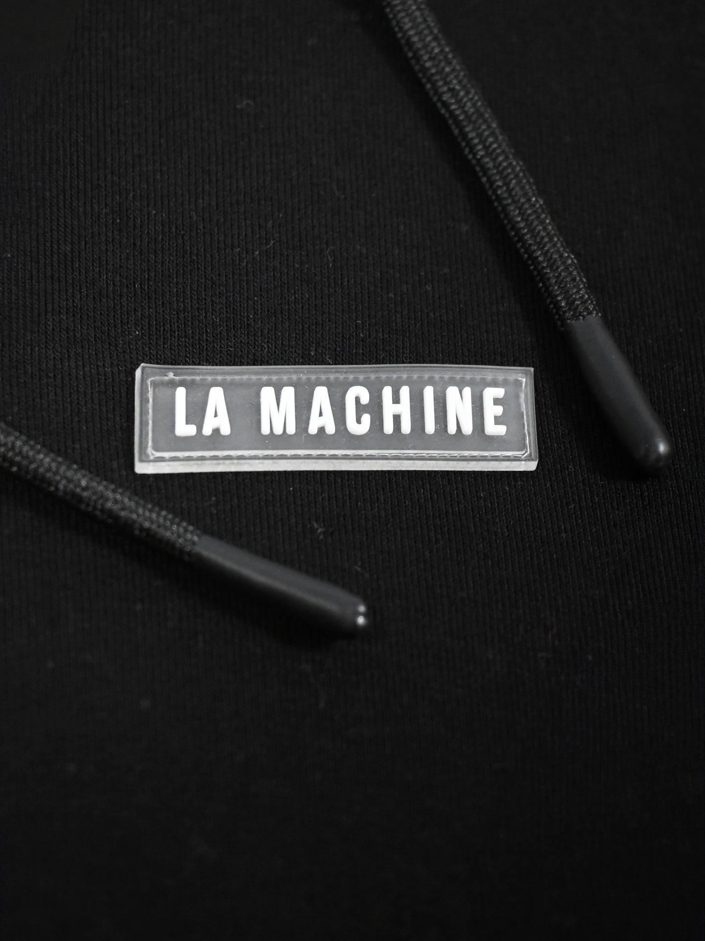Close-up of the Social Cycling Club - Hoodie by La Machine Cycle Club: a black unisex sweatshirt with two black drawstrings and a bold LA MACHINE rectangular label, perfect for cycling or showing your club pride.