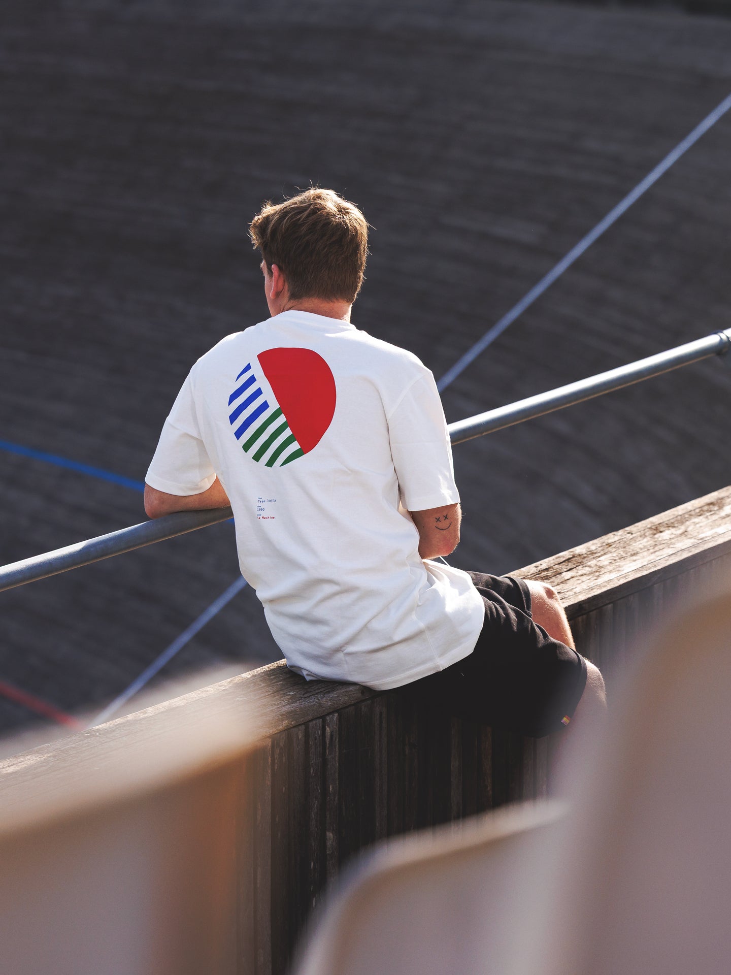 A person in a La Machine Cycle Club Team Toshiba 90 Loose Fit T-shirt with a bold red circle and diagonal stripes sits on a wooden railing, facing away and overlooking a curved outdoor structure. The heavy cotton adds comfort to the stylish tee.