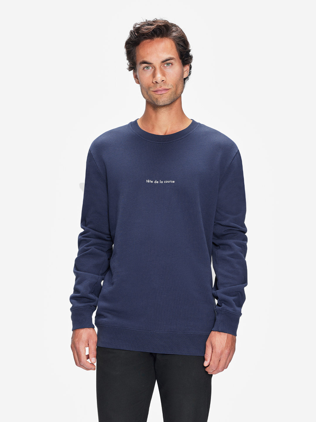 A man with short dark hair wears the La Machine Cycle Club Tête de la Course navy blue 100% cotton mens sweatshirt featuring small white île de la course text and black pants, standing against a plain white background.
