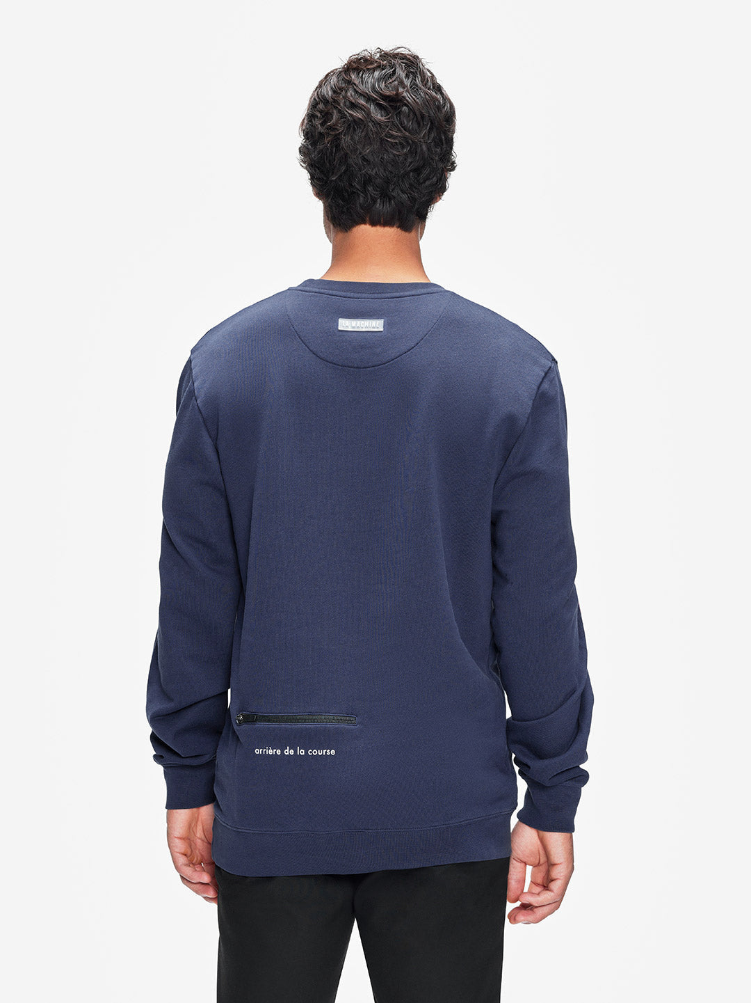A man with short, dark curly hair is seen from behind wearing the La Machine Cycle Club Tête de la Course mens navy 100% cotton sweatshirt, featuring a small white neck label and subtle lower back text in a cycling-inspired style.