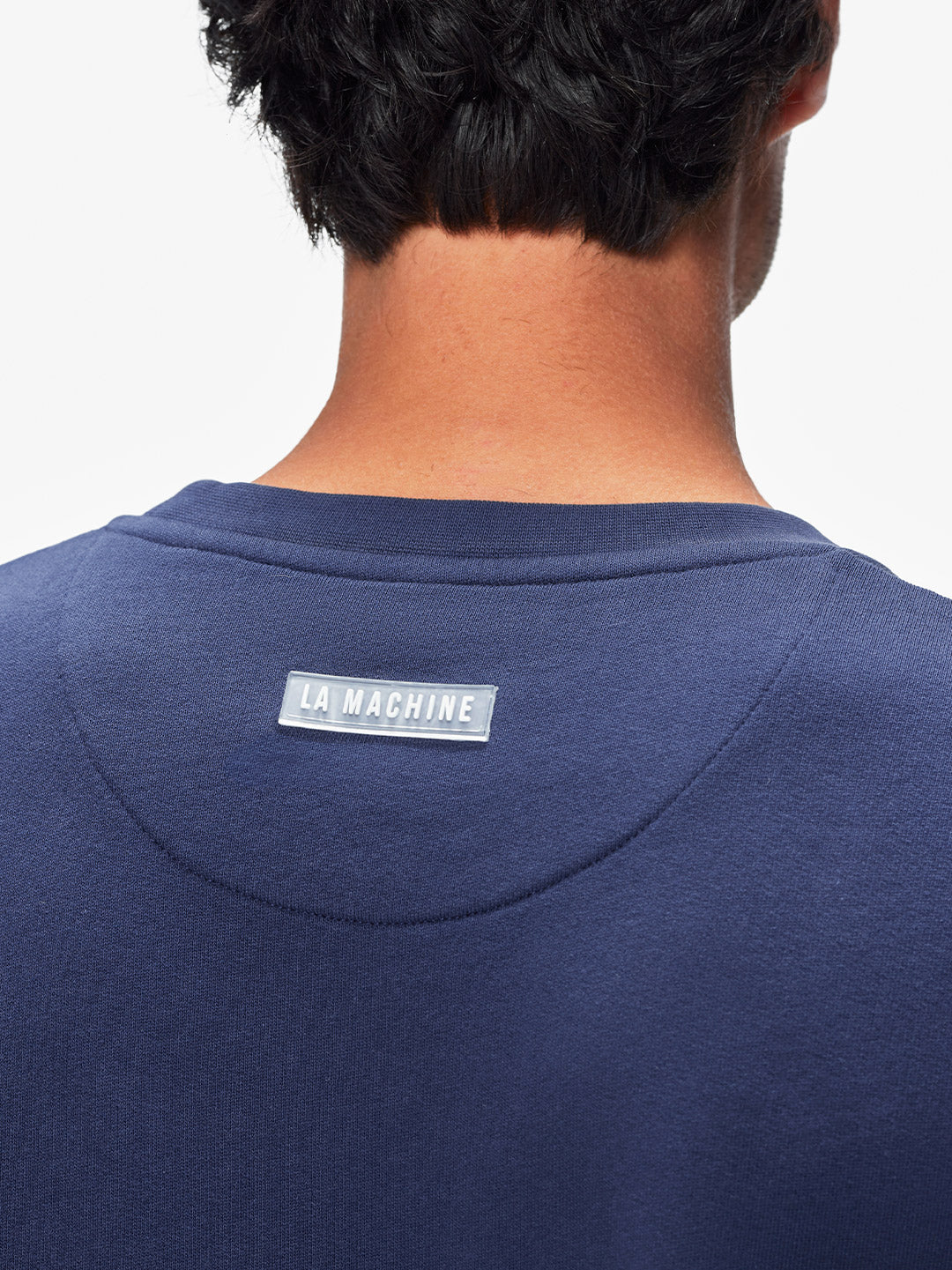 Close-up of a person’s back in the navy Tête de la Course - Sweatshirt by La Machine Cycle Club, featuring a rectangular “LA MACHINE” label near the neckline. The 100% cotton fabric and short, dark hair enhance its modern style.