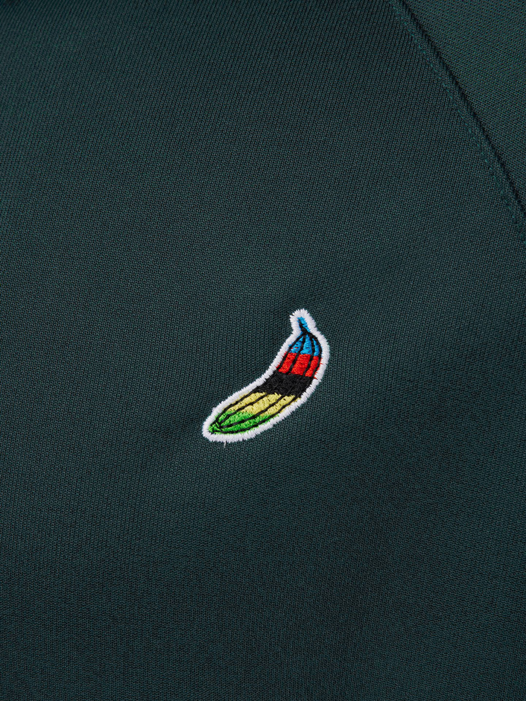 A close-up of the Atlantic Deep Food of Champions track jacket by La Machine Cycle Club shows dark green fabric and a banana embroidery in yellow, green, red, and blue outlined in white.