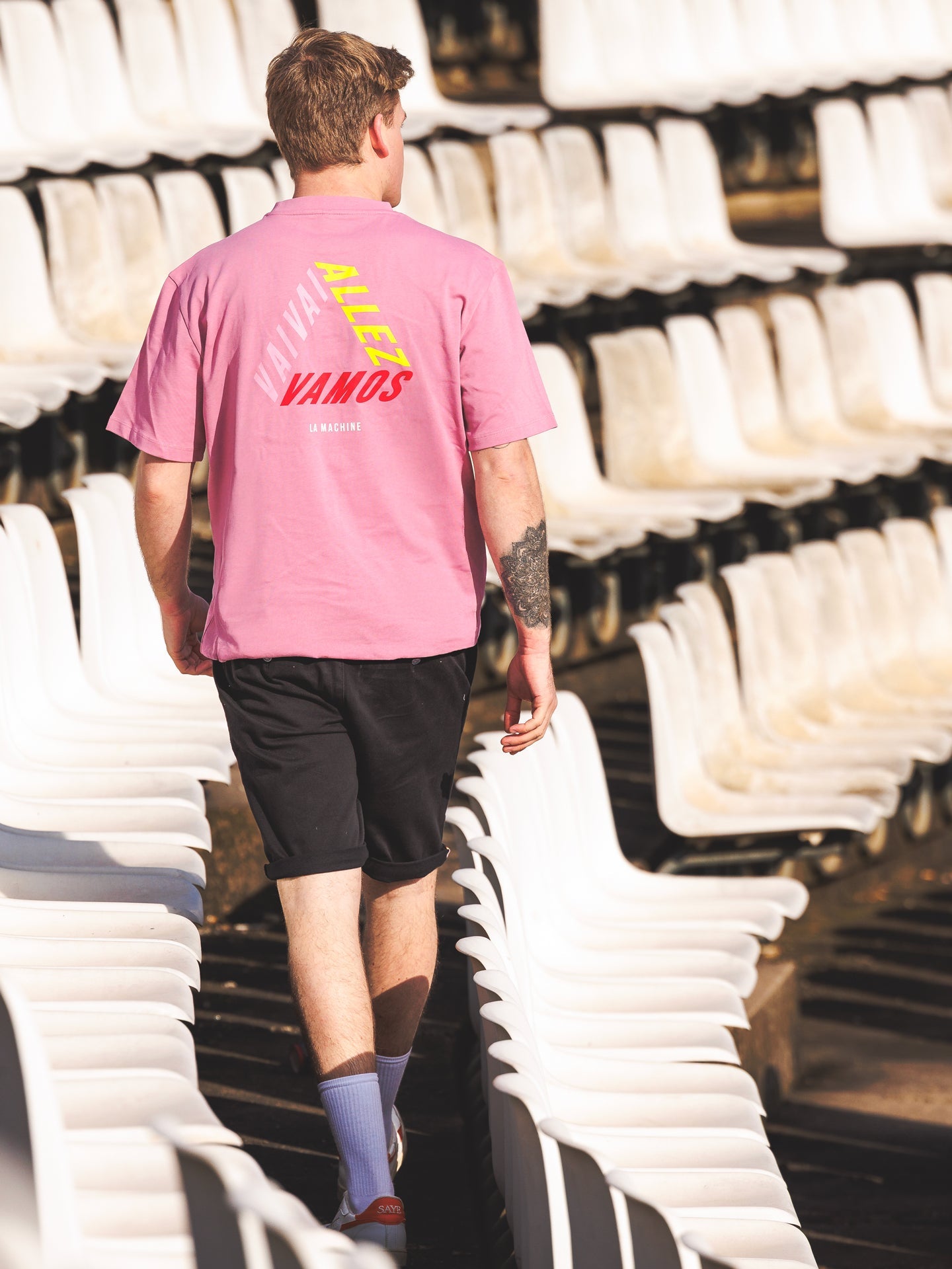 Wearing a pink Vai Allez Vamos Loose Fit T-shirt from La Machine Cycle Club, black shorts, and sneakers, a person walks through empty sunlit stadium seats. The back of this unisex cycling shirt says ALLEZ, VAMOS, and VIVA.